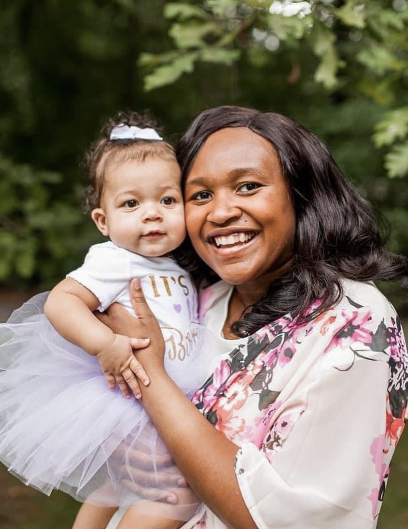 An African American woman smiling and holding a baby girl with a white bow in her hair outdoors surrounded by green foliage.