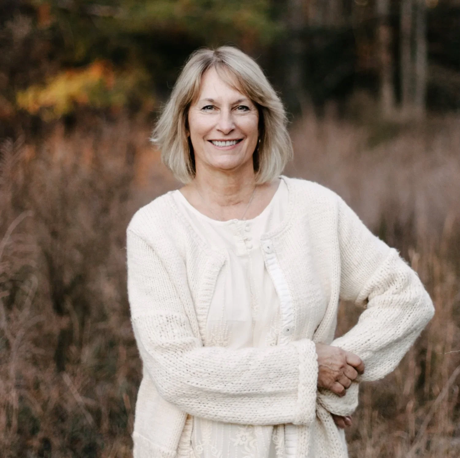 A woman with shoulder-length blonde hair, smiling, wearing a cream-colored cardigan, standing outdoors in a natural setting with tall dry grass and trees in the background.