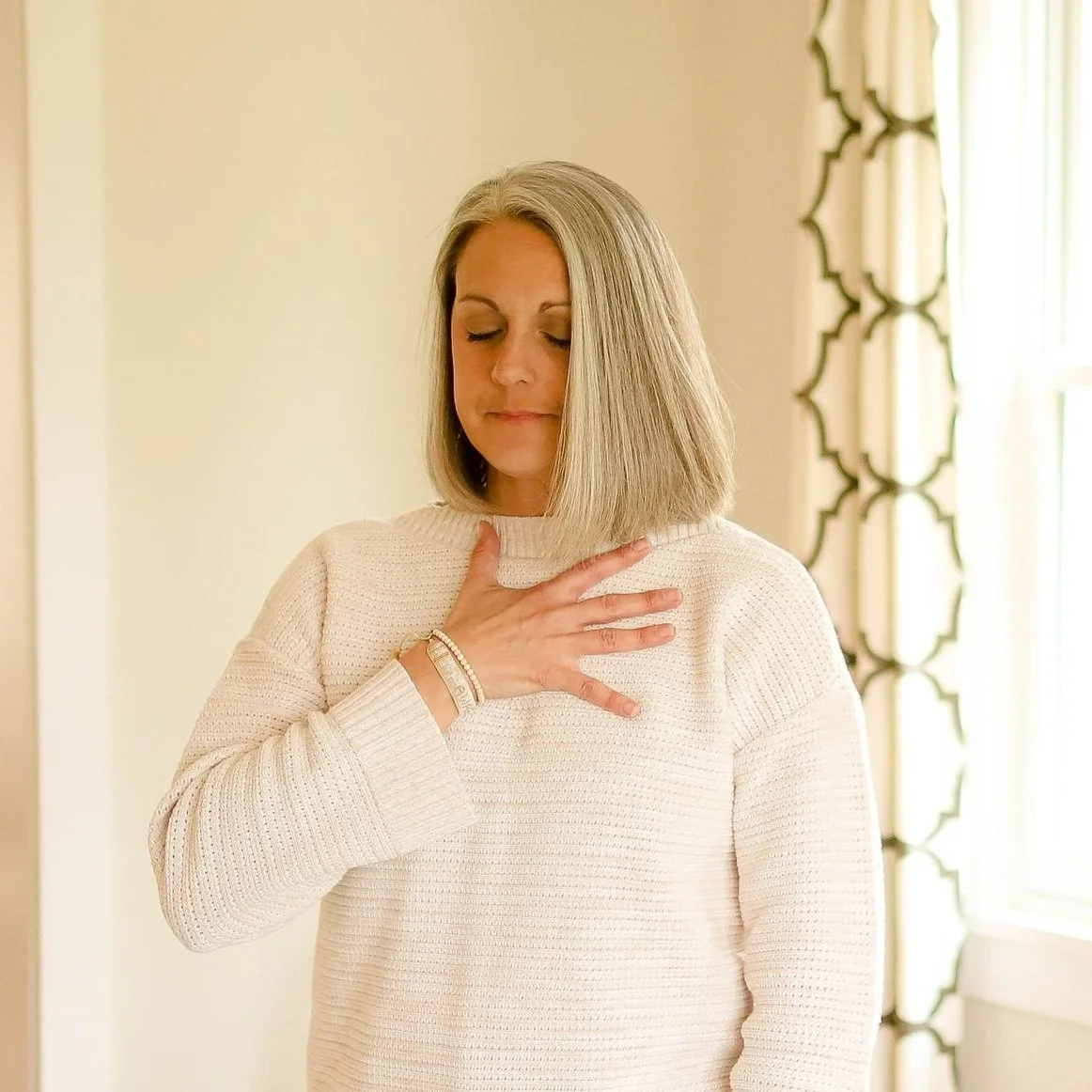 A woman with shoulder-length blonde hair placing her hand on her chest with her eyes closed in a moment of reflection or emotion, standing near a window with light-colored curtains.