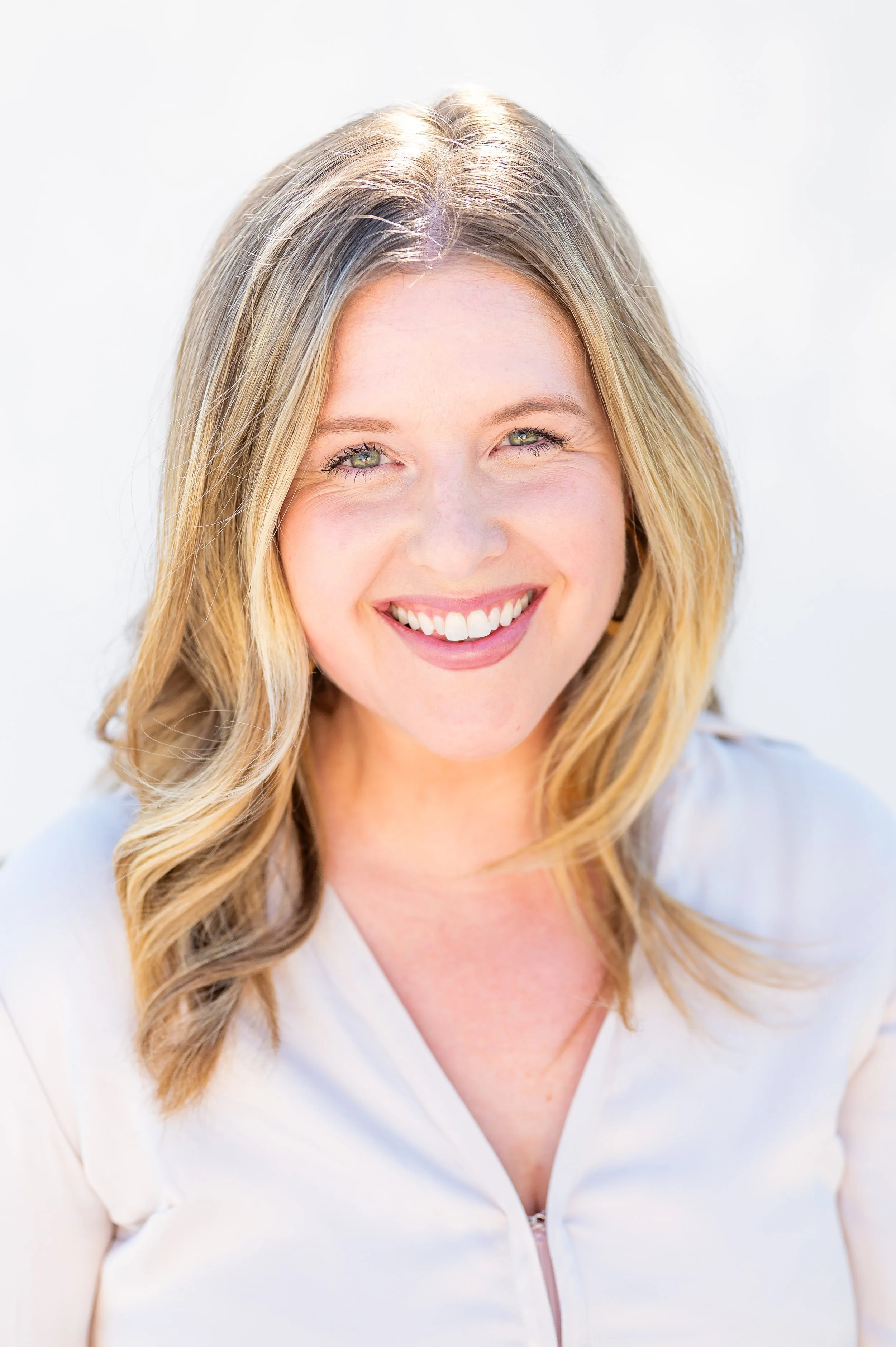 A woman with wavy blonde hair and light green eyes smiling, wearing a light-colored top, against a white background.