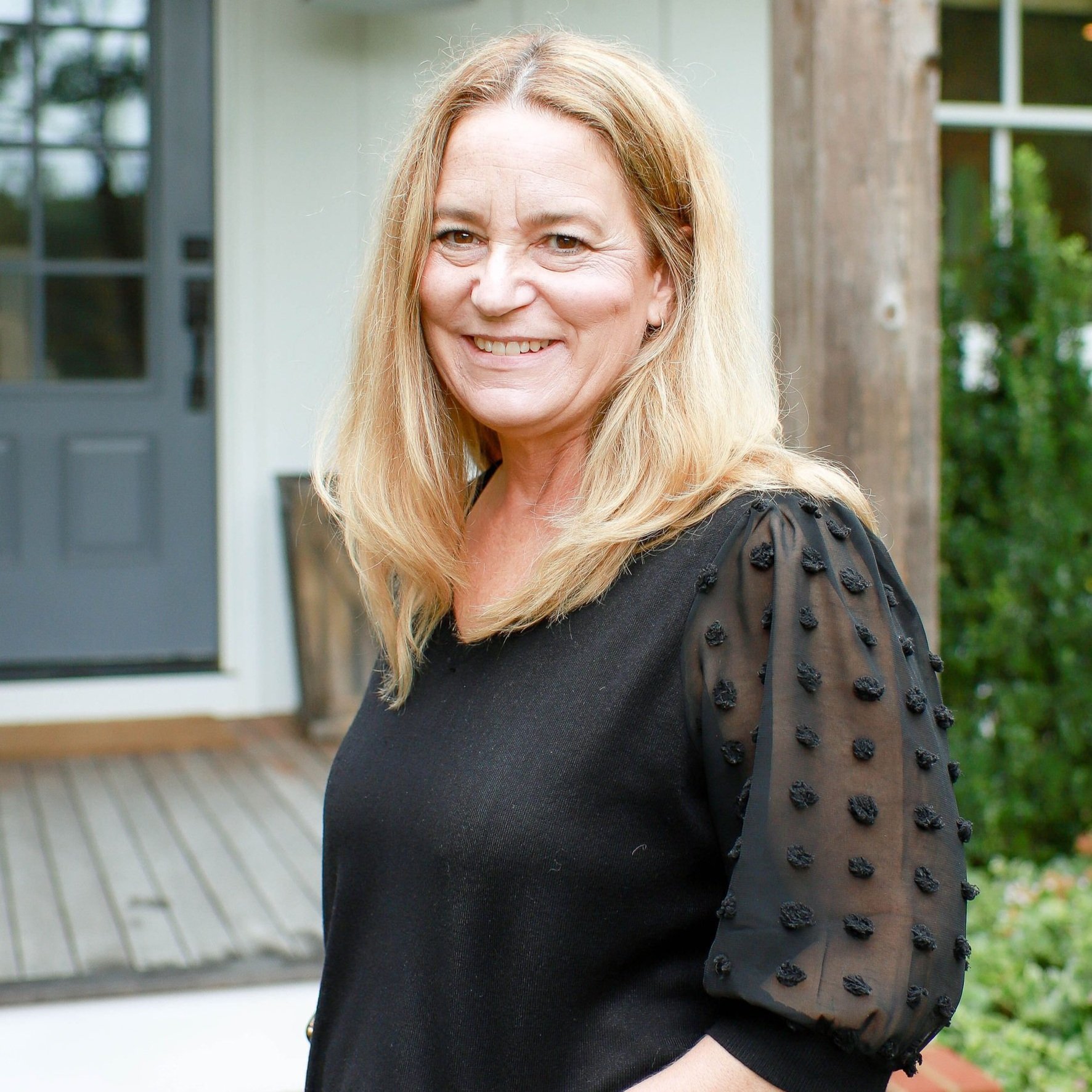 A smiling middle-aged woman with blonde hair stands outside a house, wearing a black top with sheer sleeves decorated with small black 3D bobble details.