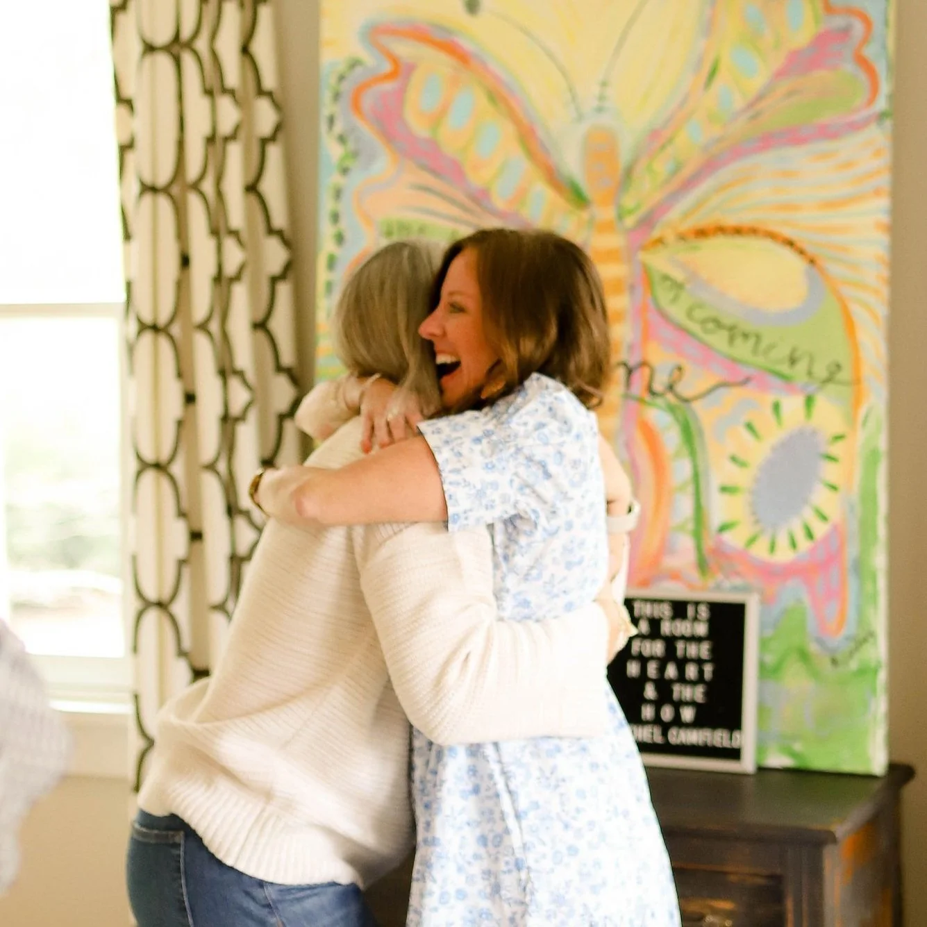 Two women hugging and smiling happily in a room decorated with bright artwork and a motivational sign.