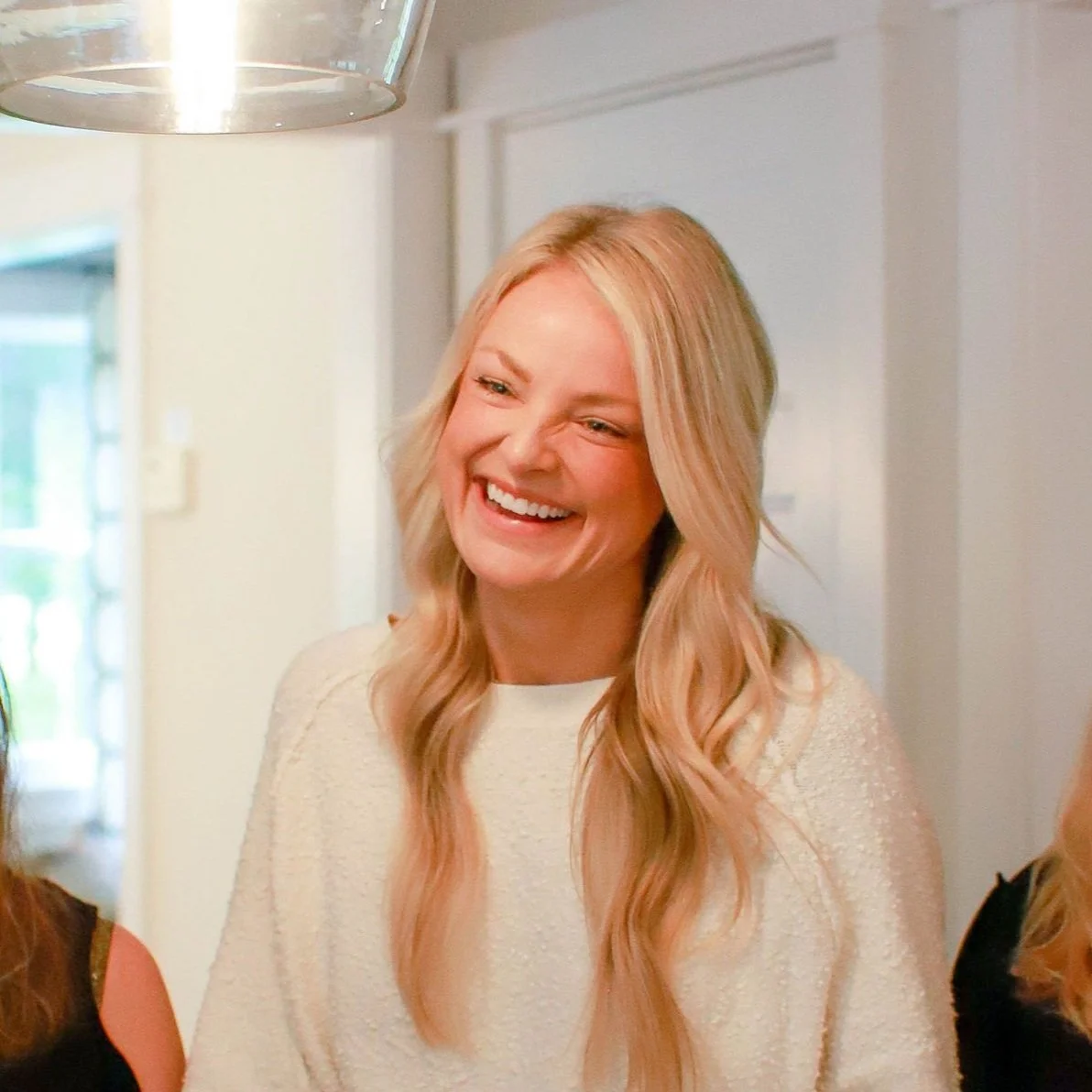 A smiling woman with long, wavy blonde hair in a white sweater indoors.