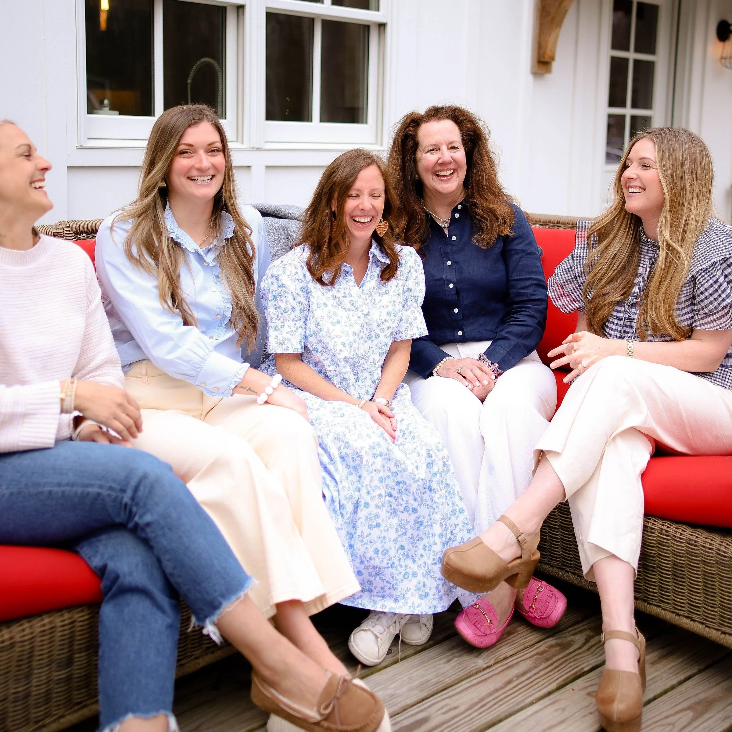 Group of six women sitting on a red outdoor sofa, smiling and laughing together, in a backyard with a wooden deck and white house in the background.