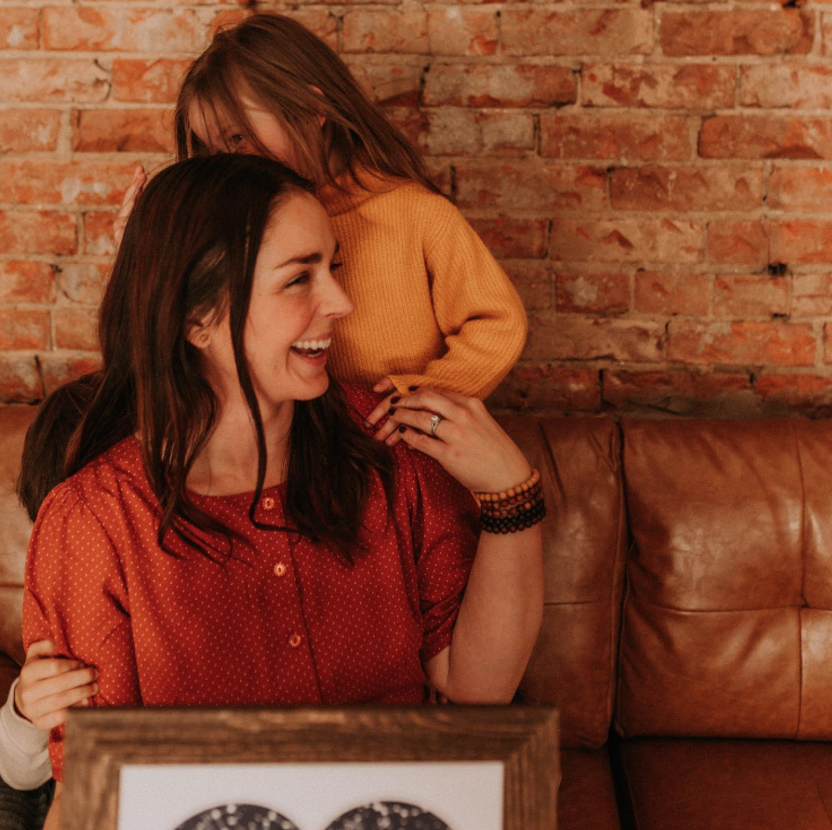 A woman and a young girl share a joyful moment inside a cozy room with an exposed brick wall. The woman is sitting on a leather couch, smiling and wearing a red patterned shirt, while the girl stands behind her with her arms around her neck, wearing a yellow sweater.