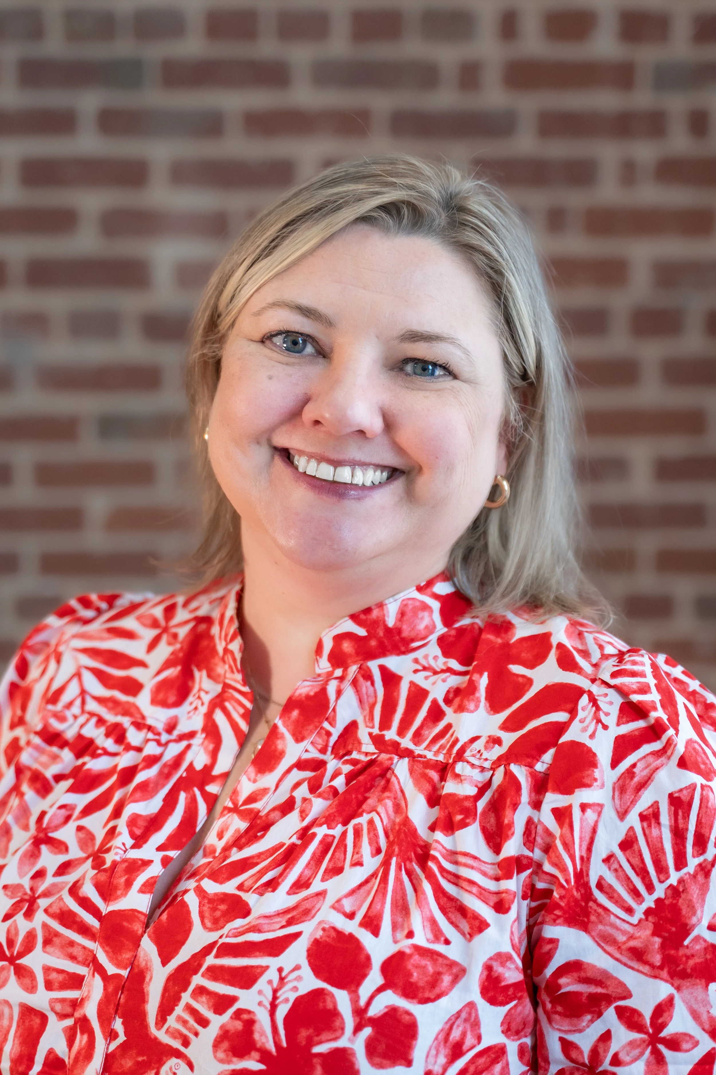 A woman with blonde hair wearing a red and white patterned shirt, smiling, with a brick wall background. Key Goforth, Director of Elementary Programs and Interventions at Gainesville City Schools. 