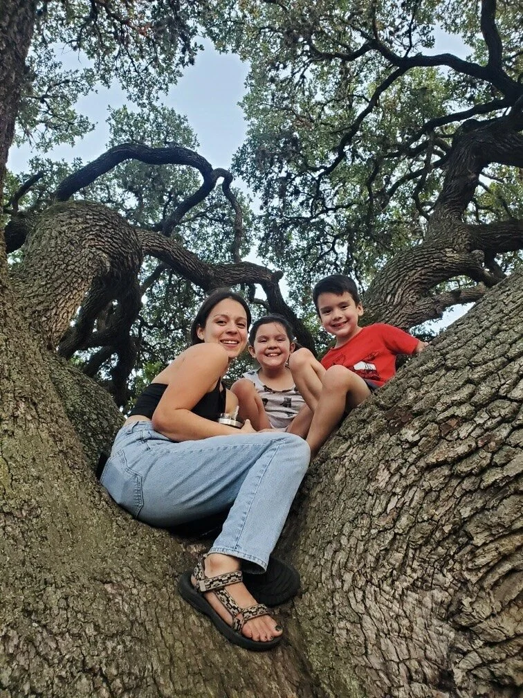 A woman and two children sitting in the branches of a large tree with green leaves, smiling at the camera.