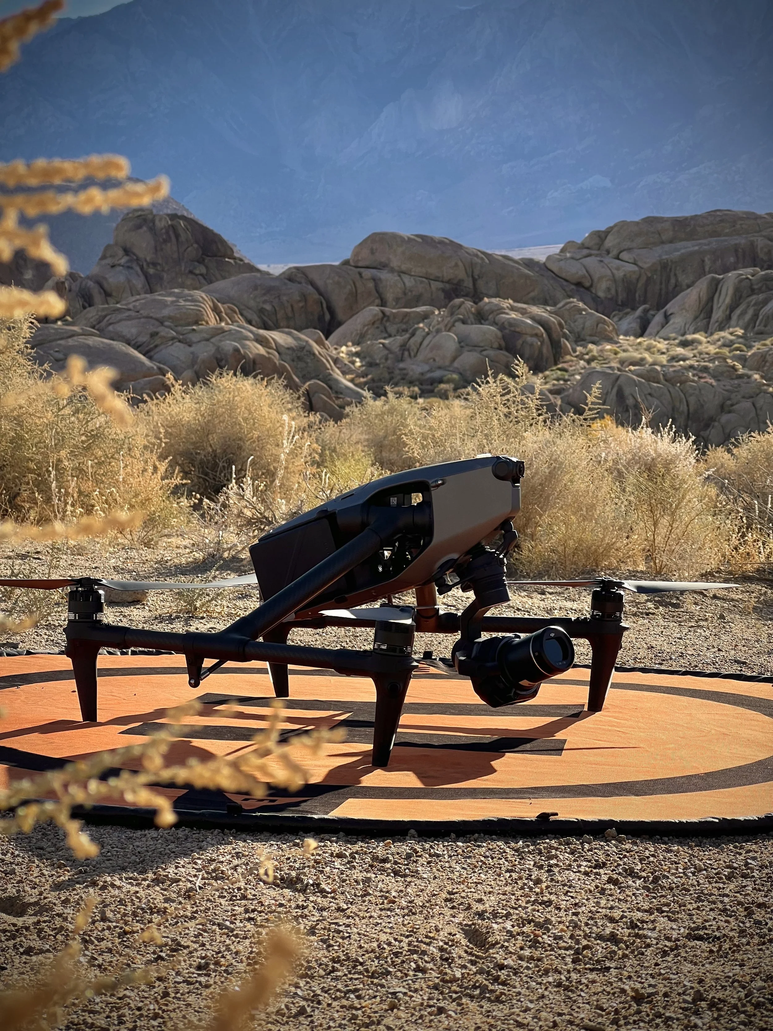 A drone is placed on an orange landing pad in a desert landscape with rocks and sparse bushes, mountains in the background, under a clear blue sky.