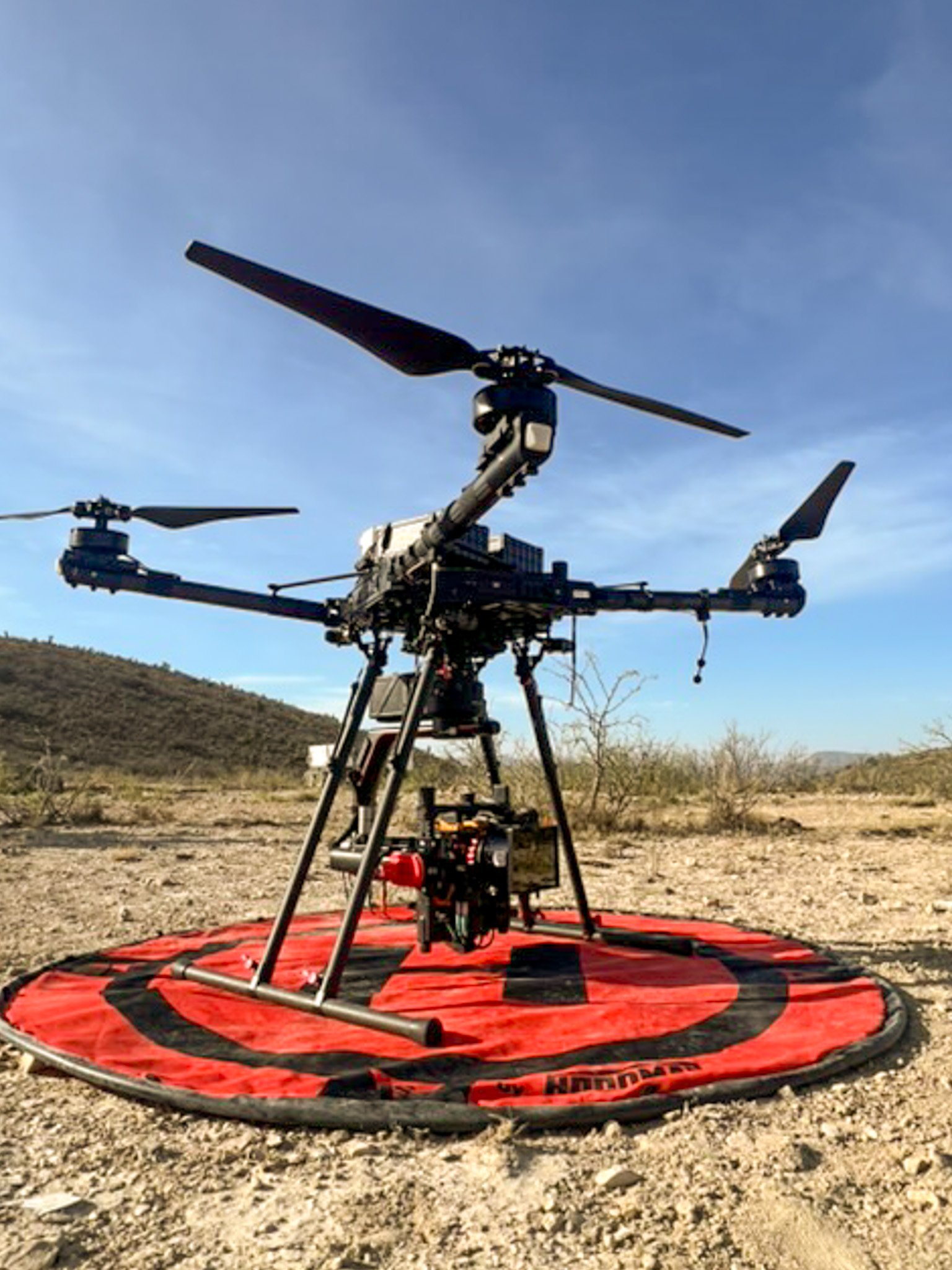 A large drone with four rotors, standing on a red and black landing pad in a desert landscape with hills in the background.