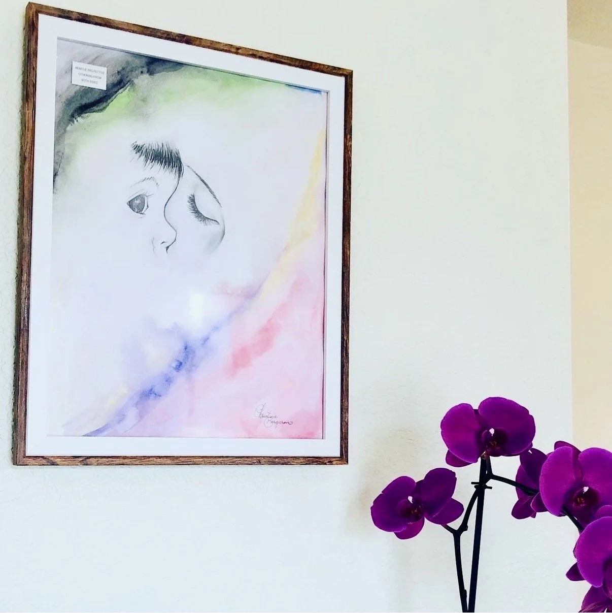 A framed watercolor painting of a woman's face with closed eyes and long eyelashes, on a light-colored wall. There are purple orchids in the foreground on the right side of the image.