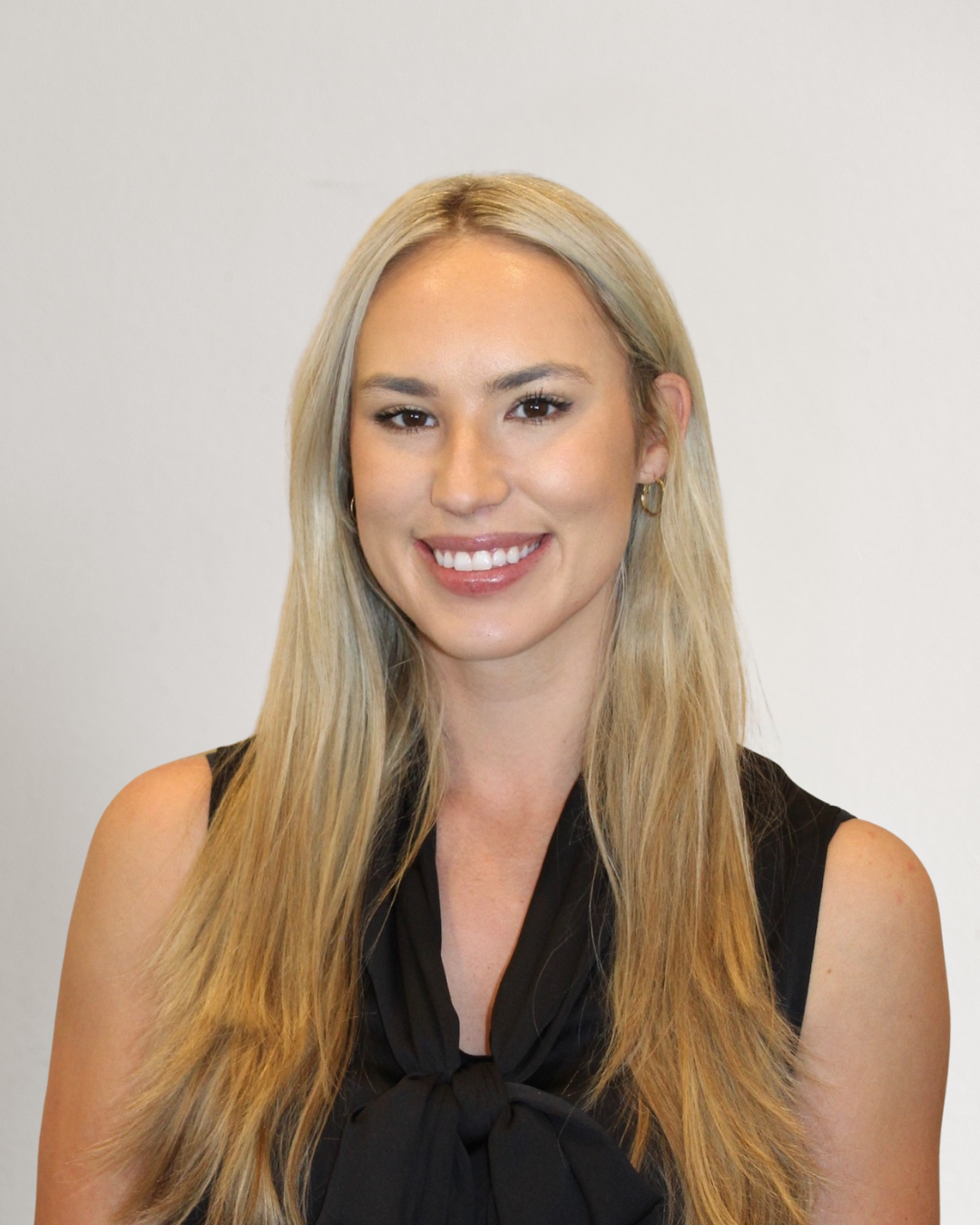 A smiling woman with long blonde hair, wearing a sleeveless black top with a bow tie, standing in front of a light-colored background.
