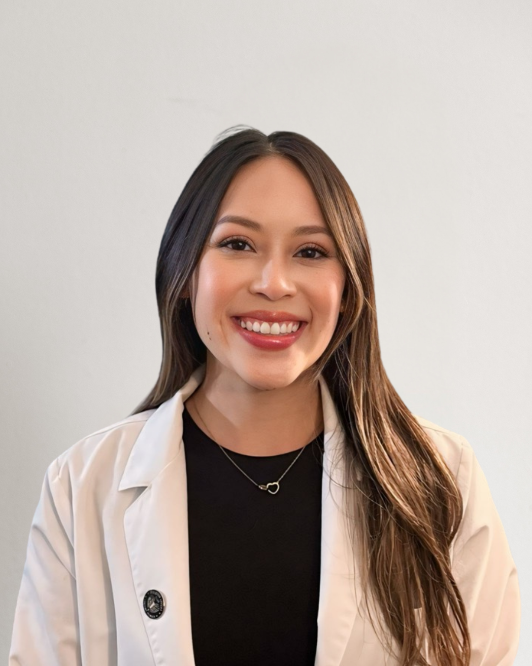A young woman with long brown hair, smiling, wearing a white coat over a black top, and a delicate necklace, standing in front of a beige background.