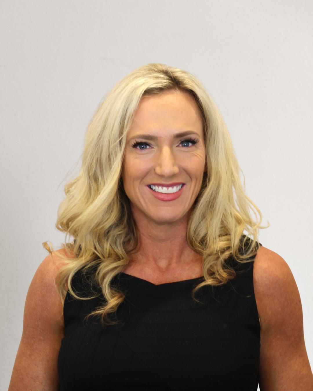 A woman with long, curly blonde hair, wearing a black sleeveless top, smiling against a light-colored background.