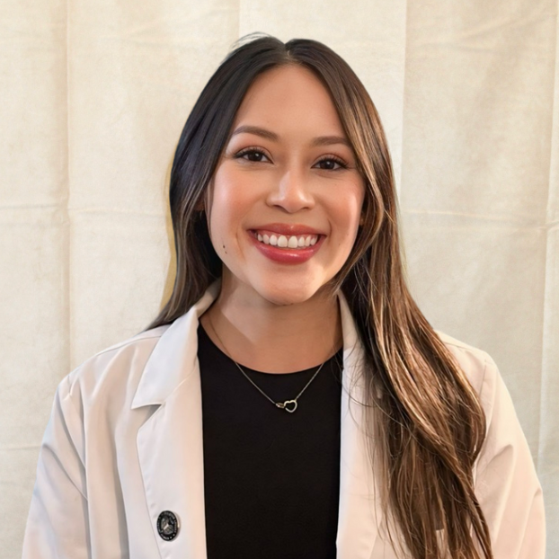 A young woman with long brown hair, smiling, wearing a white coat over a black top, and a delicate necklace, standing in front of a beige background.