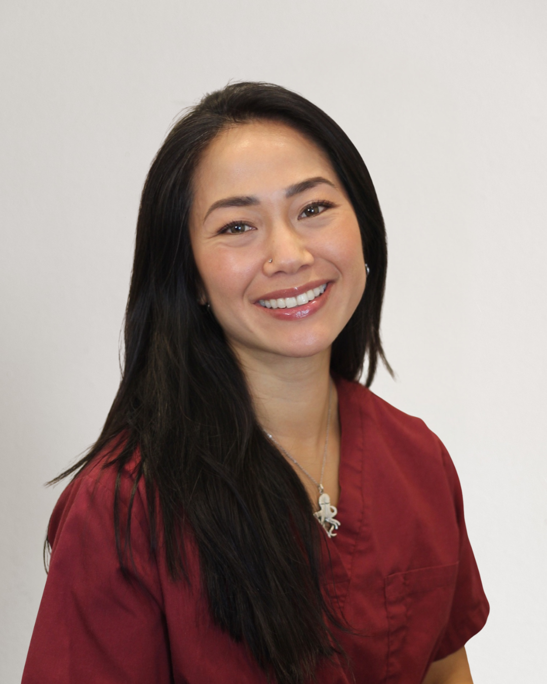 A woman with long black hair wearing a red medical uniform, smiling, standing against a neutral background.