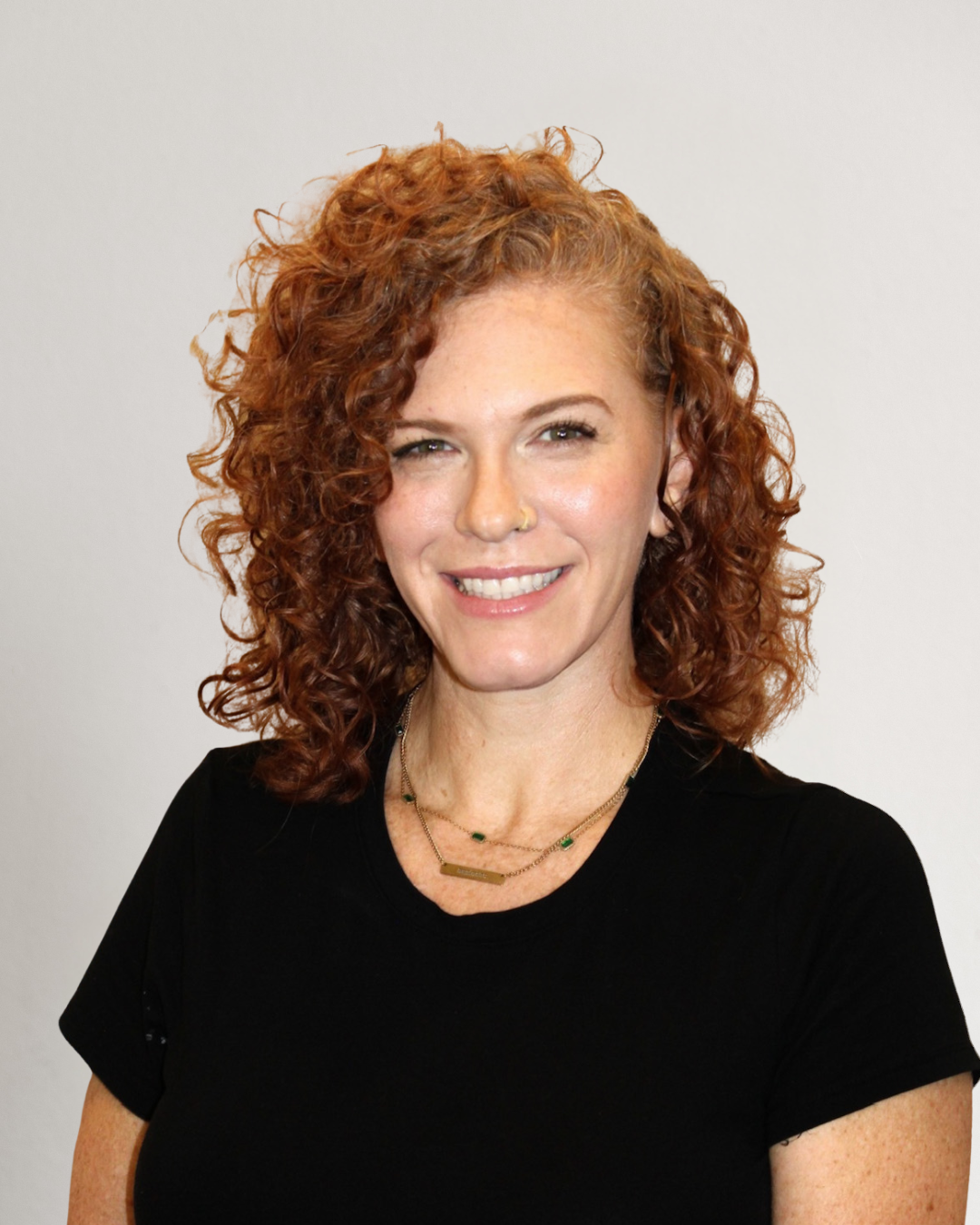 A woman with curly red hair, fair skin, and freckles, smiling, wearing a black t-shirt and layered necklaces, standing in front of a beige curtain.