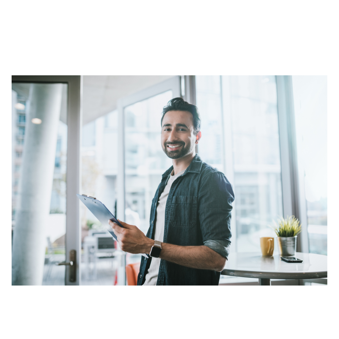 Smiling man holding clipboard in modern office with large windows and plant on table.