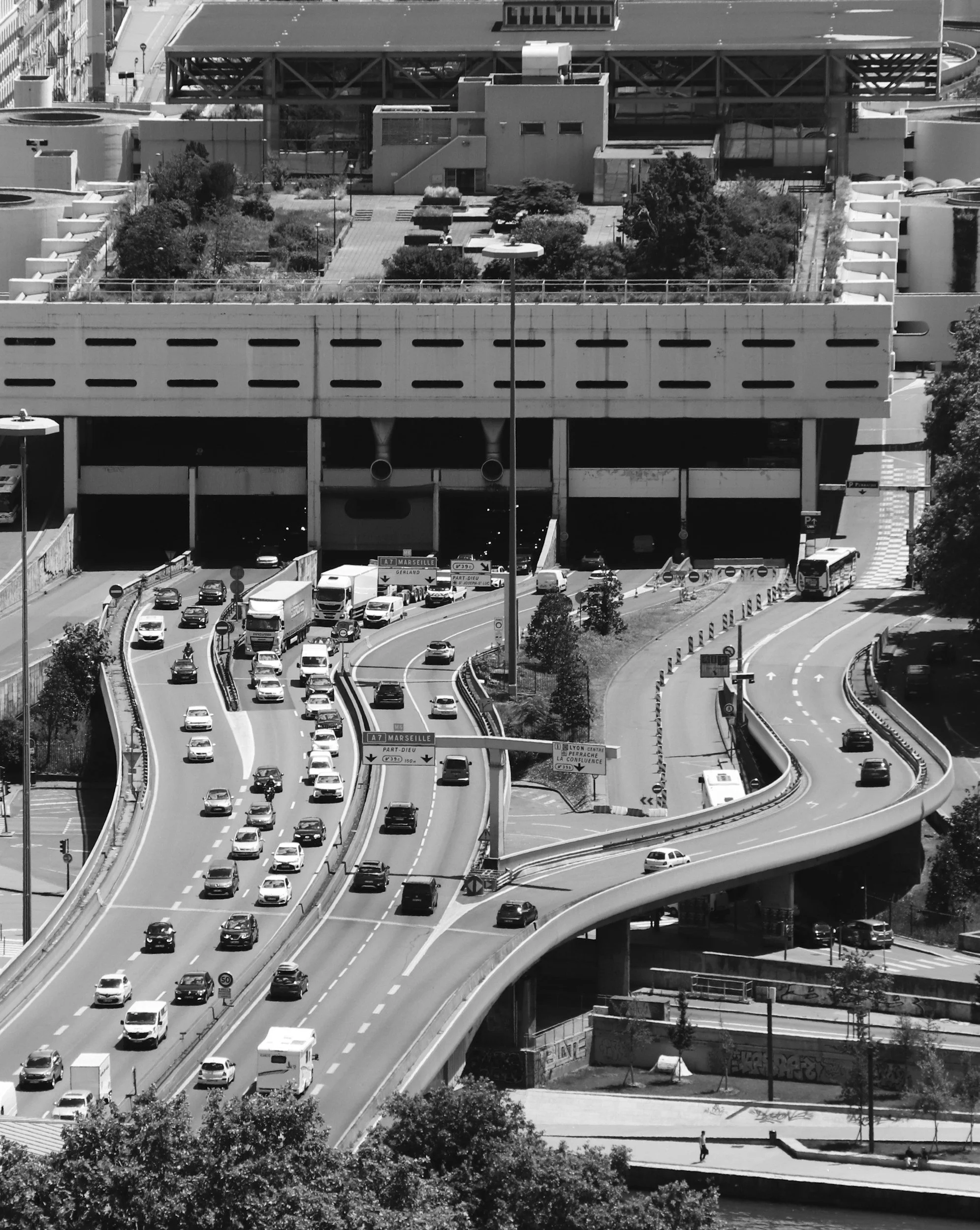 A busy multi-level highway with cars and trucks, overpasses, and a modern building with trees and pedestrians nearby.