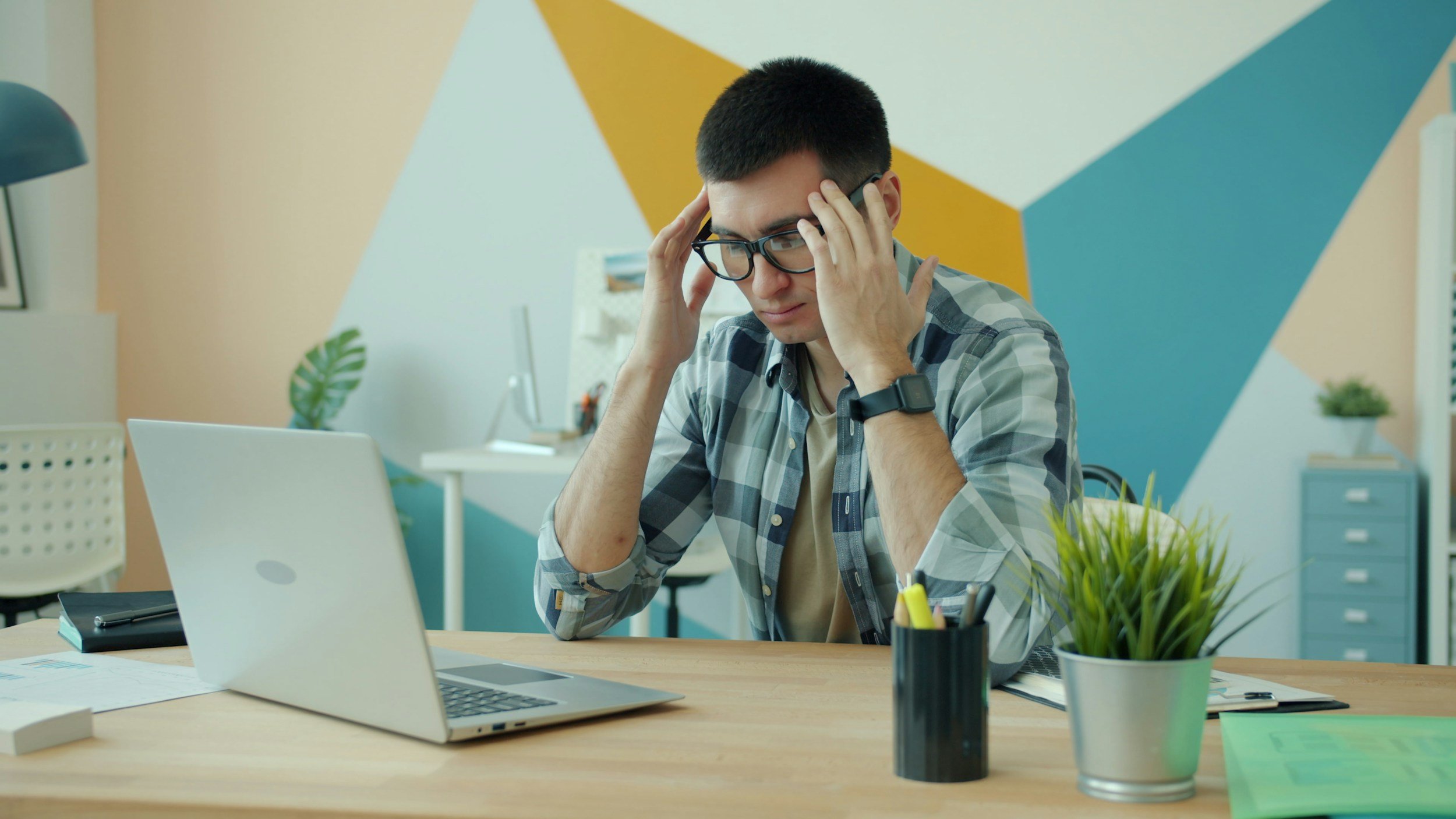 Young man with glasses and a plaid shirt sitting at a desk with a laptop, looking stressed or confused, in a colorful modern office.