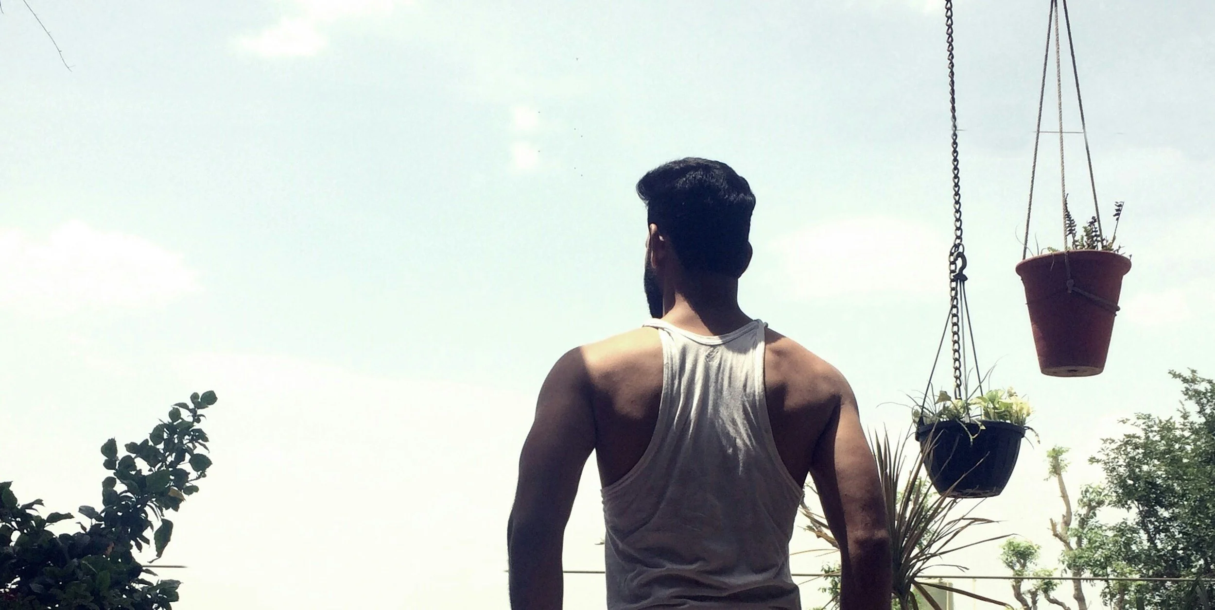 A man with dark hair and a beard, seen from behind, stands outdoors on a bright day, wearing a white tank top, with plants and hanging flower pots nearby.