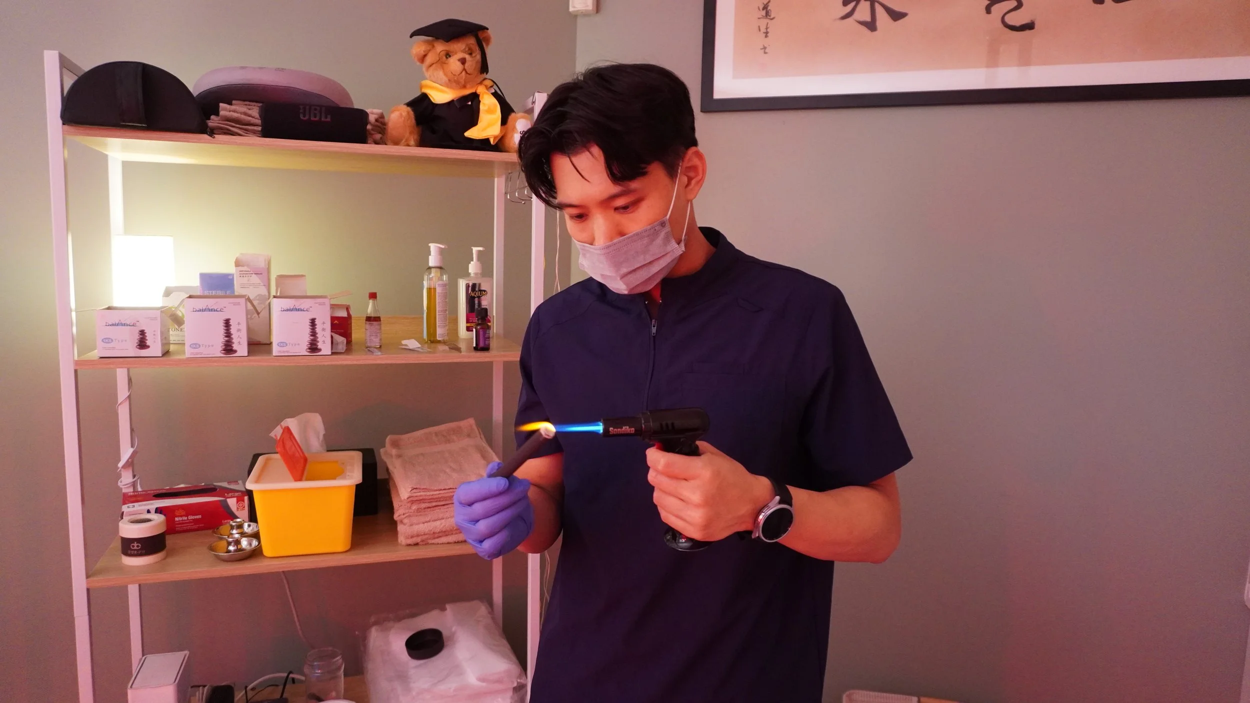 A man wearing a face mask and gloves uses a blowtorch on a small object in a medical or lab setting.