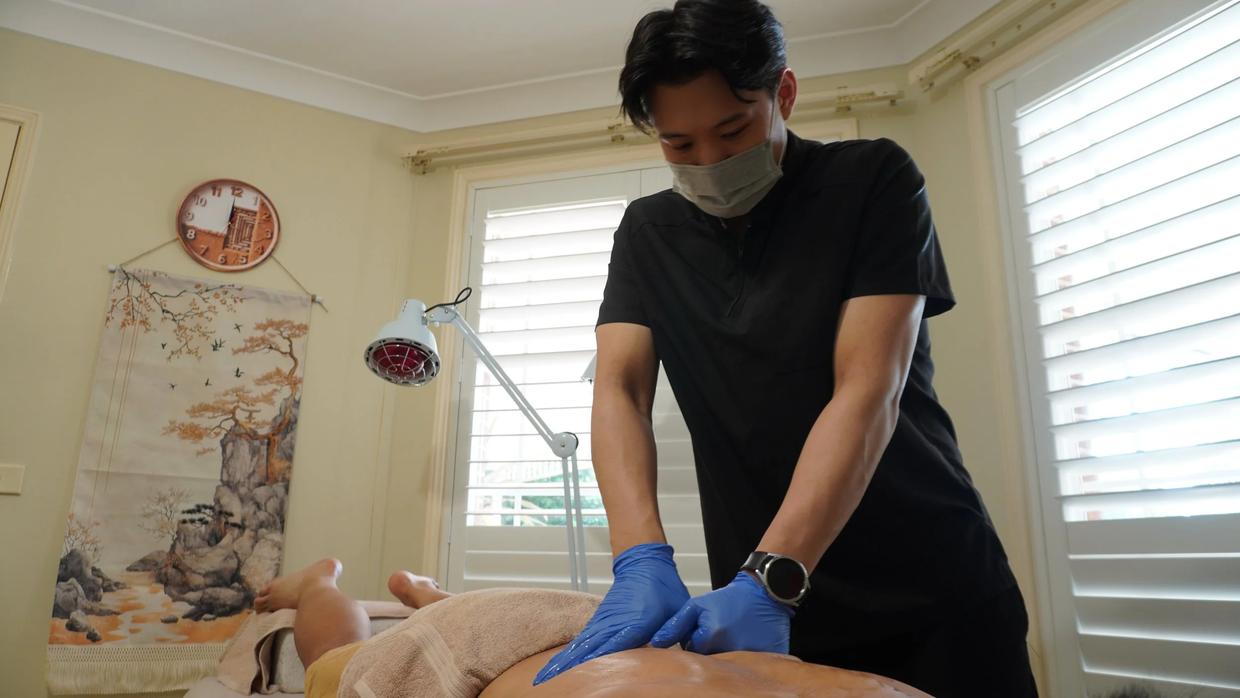 A massage therapist wearing a face mask and blue gloves performing a massage on a client lying face down on a massage table, in a room with white shutters and a wall clock.
