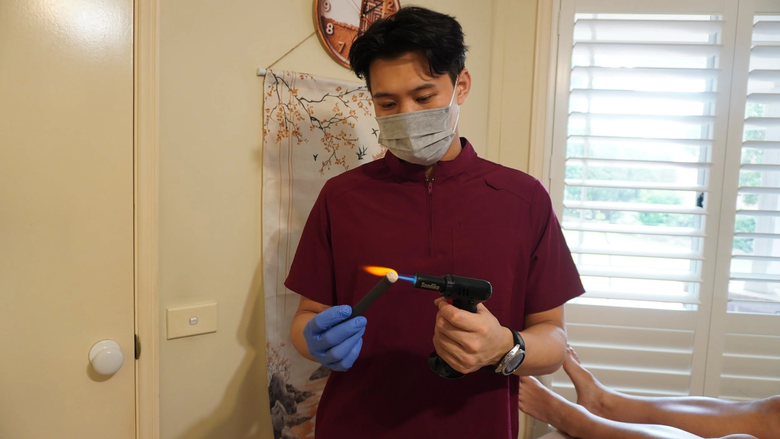 A medical professional with black hair, wearing a gray face mask, blue gloves, and a maroon scrubs, is holding a lit soldering iron and a stick of material in a room with cream-colored walls, a wall clock, industrial shutters, and traditional Asian d