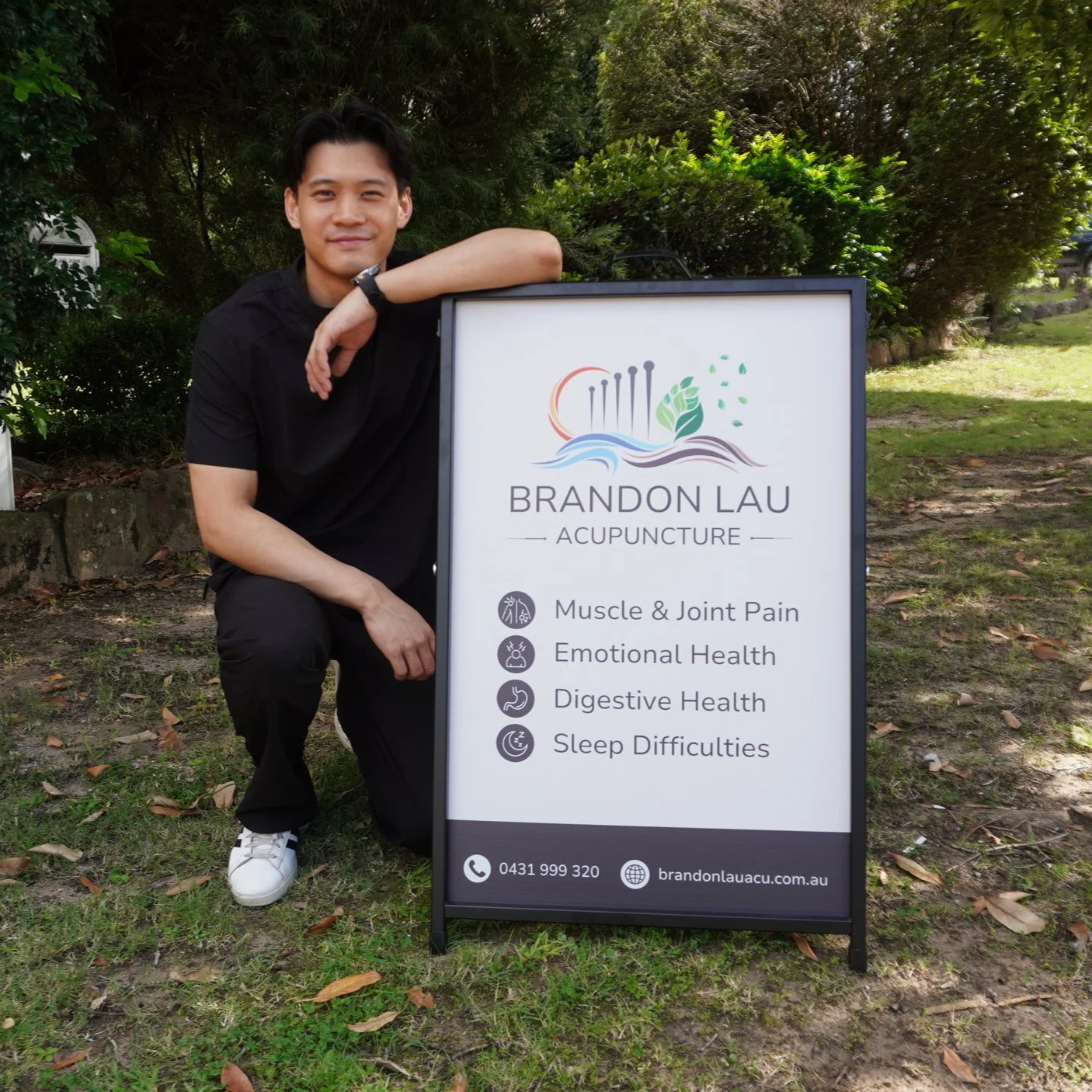 Young man kneeling next to a sign for Brandon Lau Acupuncture, with tree and grass background.