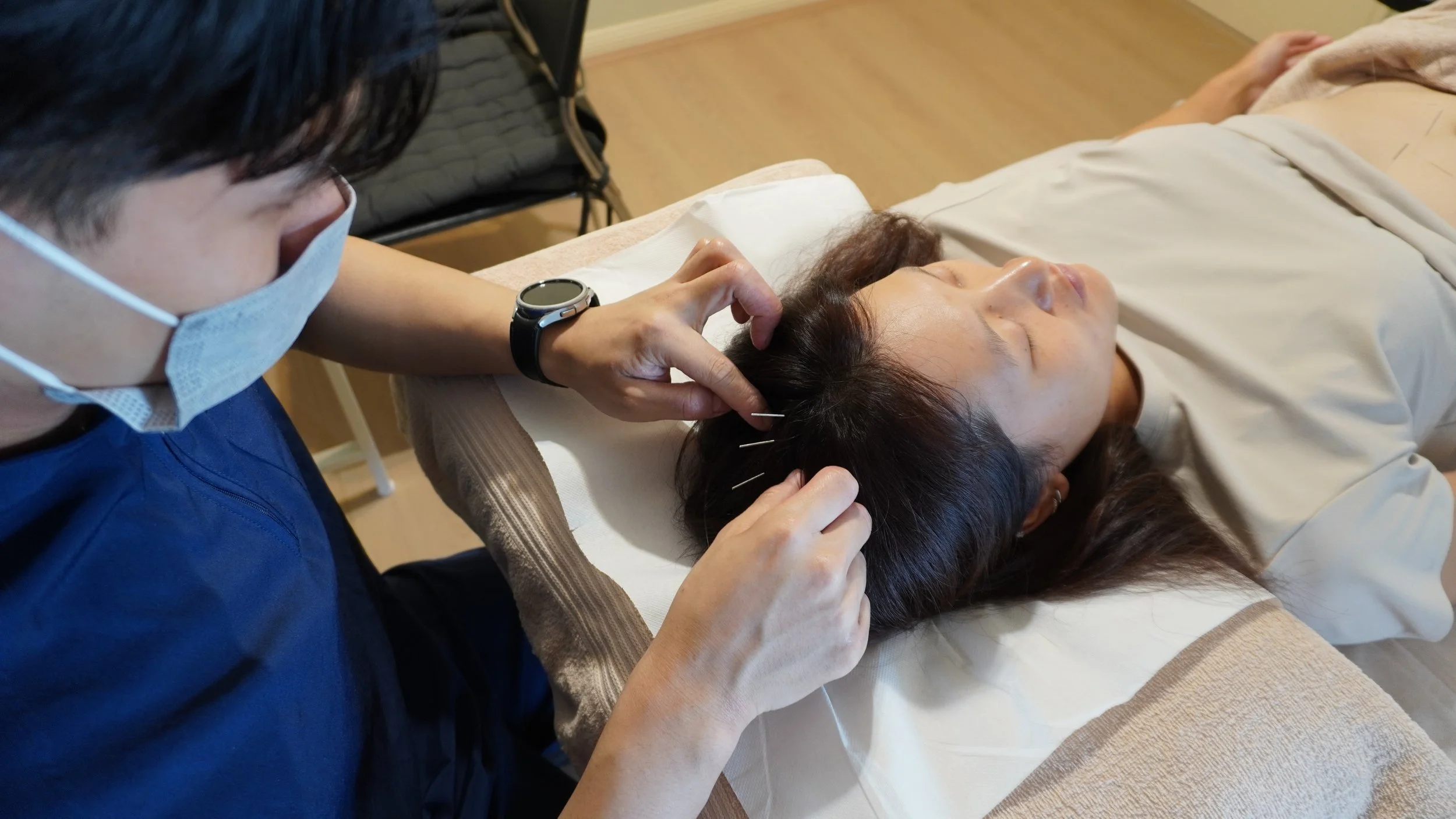 A person receiving acupuncture treatment on their scalp from a practitioner wearing a face mask.