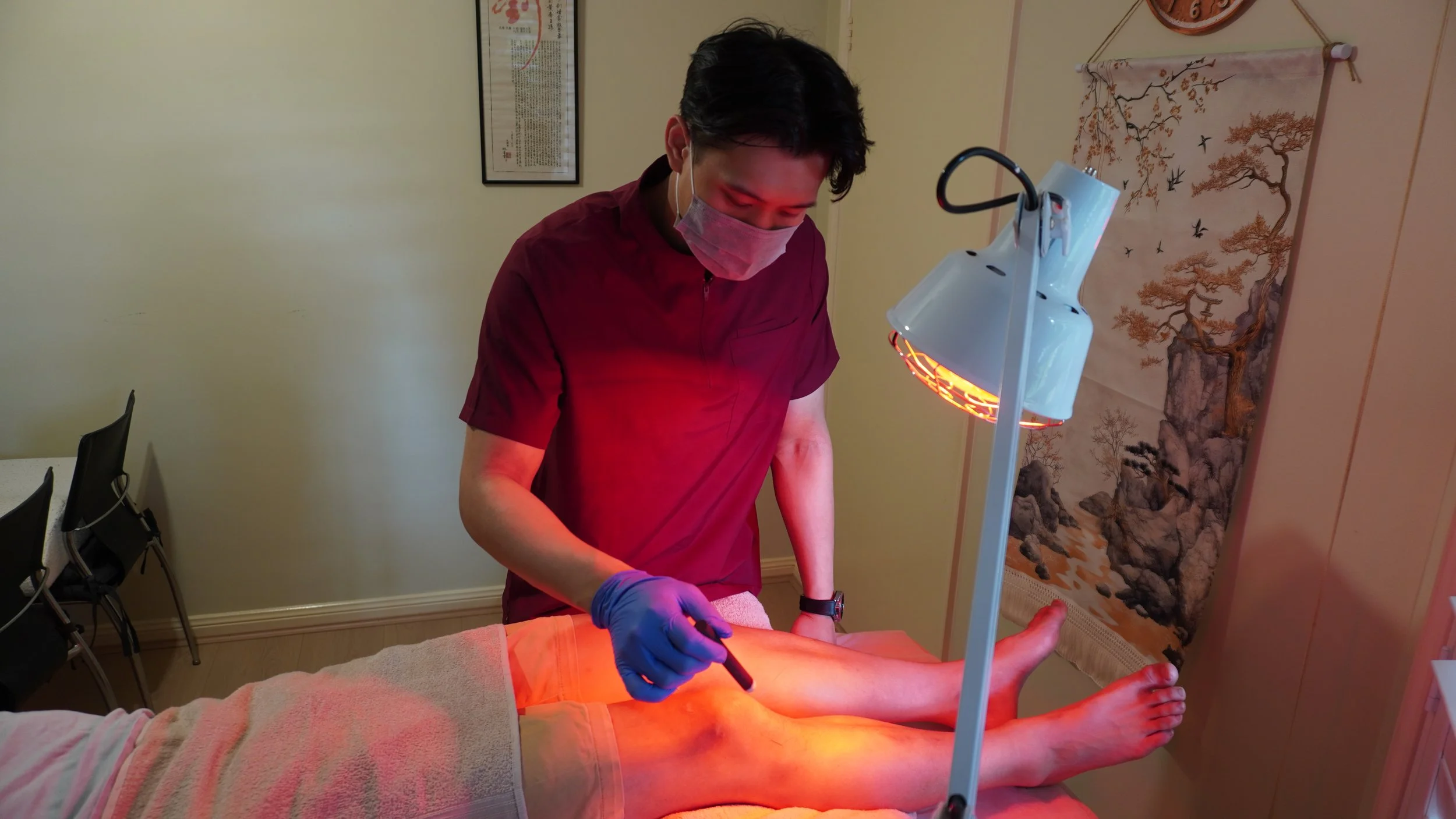 A person receiving a foot treatment or therapy under a heat lamp, with a practitioner applying a device to the person's foot, in an indoor setting with Asian art on the wall.