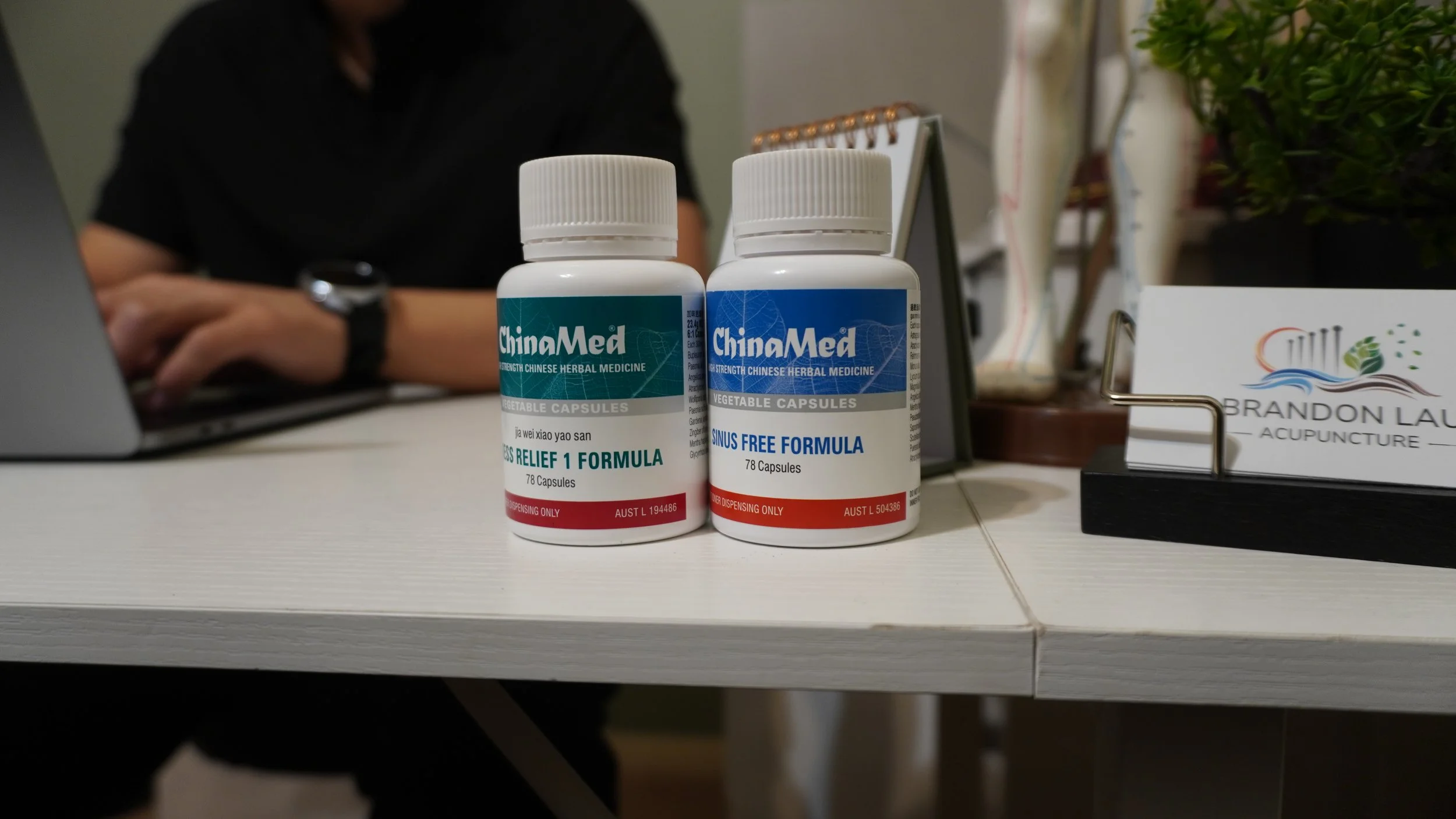 Two bottles of Chinese herbal medicine capsules on a table, labeled 'Relief 1 Formula' and 'Sinus Free Formula,' with a person seated in the background working on a laptop and a business card for acupuncture services nearby.