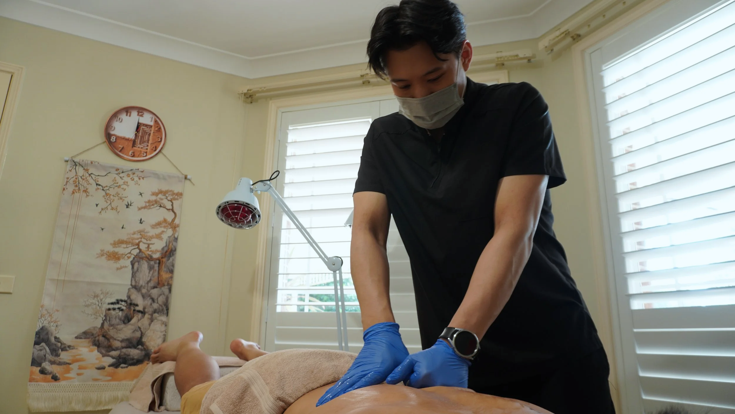 Therapist providing physical therapy to a patient lying on a table, wearing gloves and a mask, in a relaxed room with a large window and traditional wall art.
