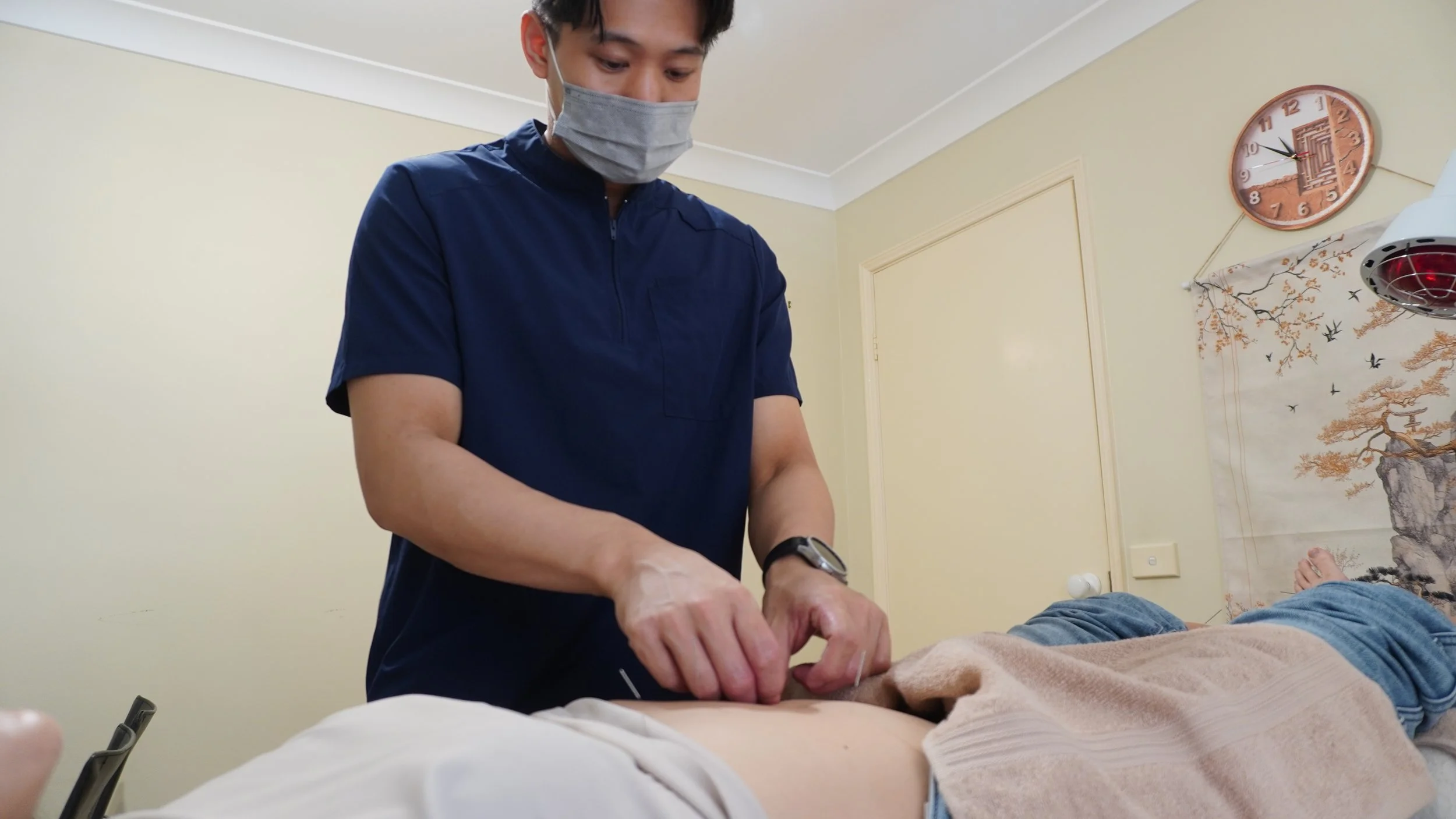 A healthcare professional in a grey face mask and dark blue scrubs performs acupuncture on a patient lying on a medical bed in a clinic room.