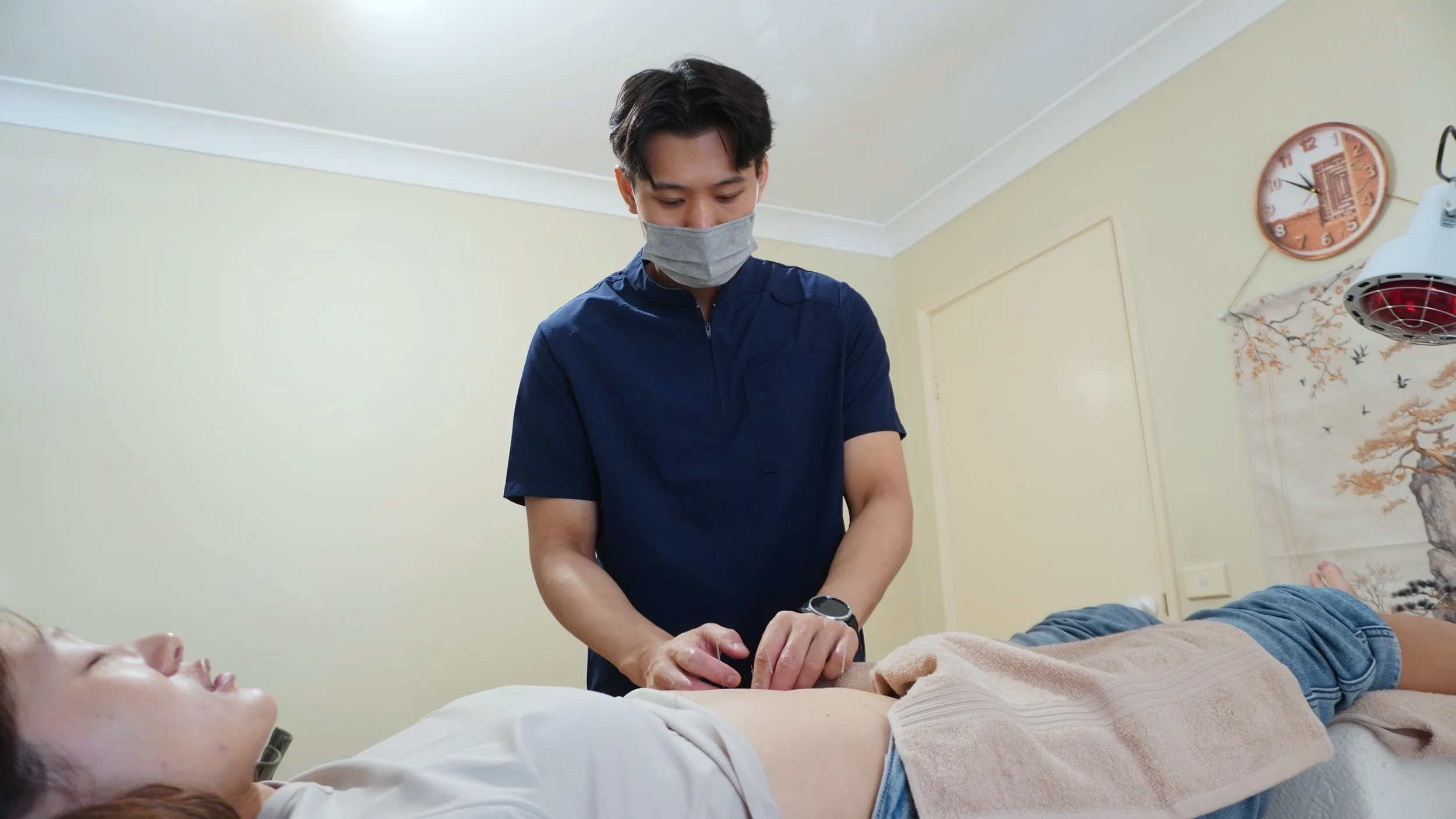 A healthcare professional performing a medical procedure on a patient's abdomen in a clinical room with a decorative wall art and wall clock.