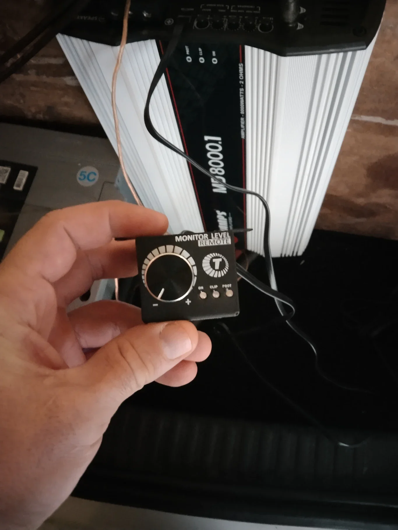 Une main tenant un contrôleur à niveau de moniteur à distance noir, avec des boutons et un grand cadran, devant une grosse batterie ou un amplificateur.