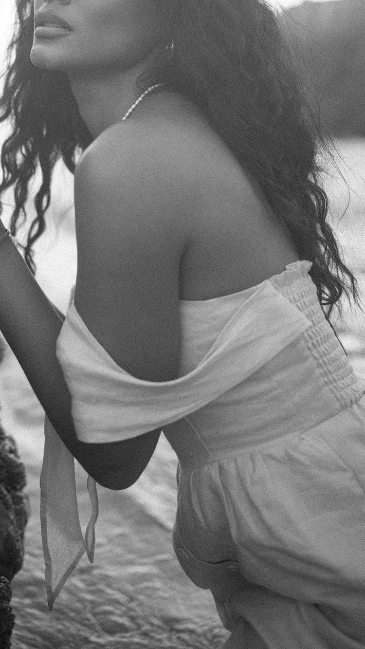 Close-up van een vrouw met golvend haar, zittend op het strand, in het zwart-wit, met een parelsnoer en een off-shoulder top.