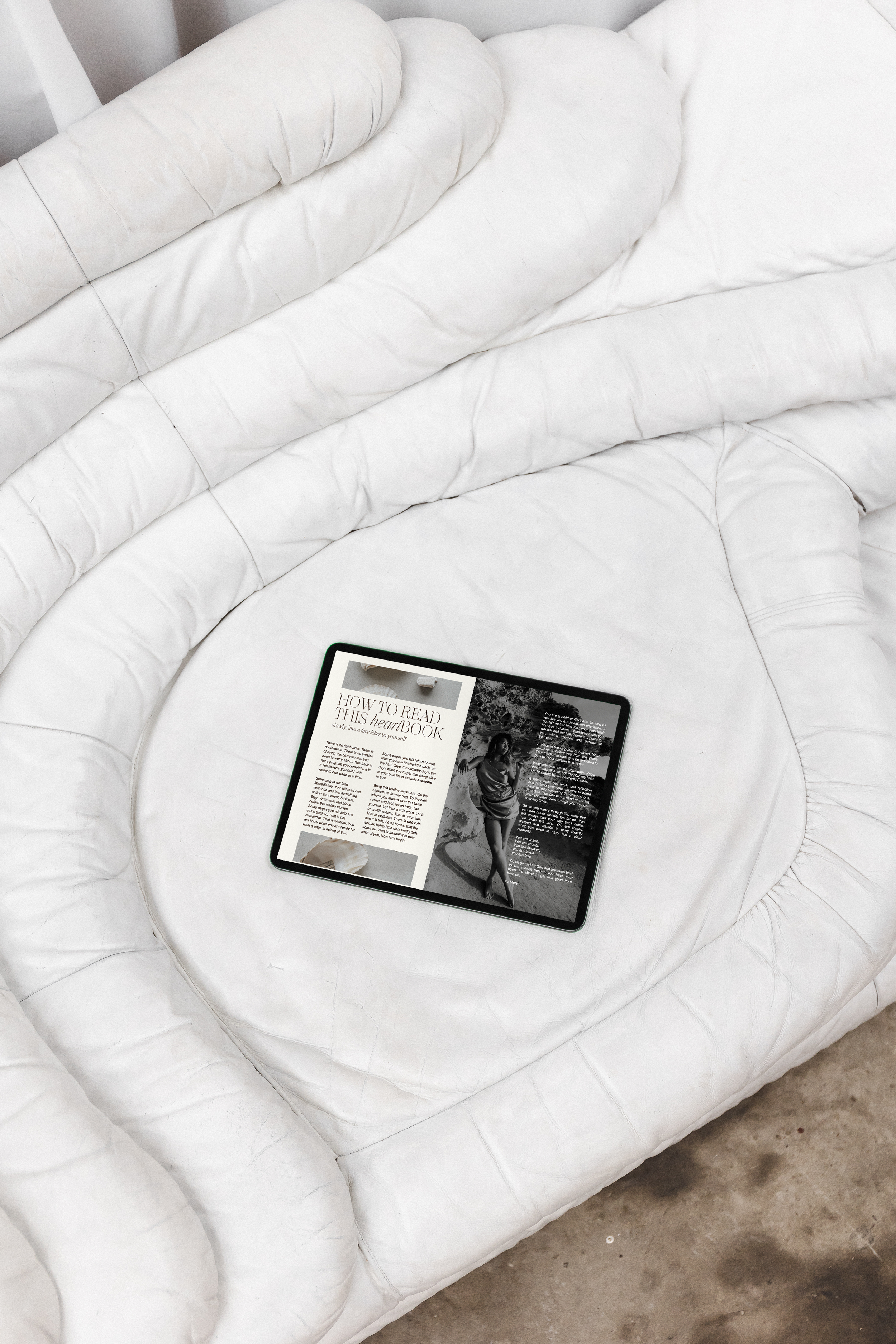 Een wit, comfortabel uitziend bankje of fauteuil met een tablet erop dat een artikel toont met een zwart-wit foto van een vrouw in een jurk. De bank staat op een cementen vloer.