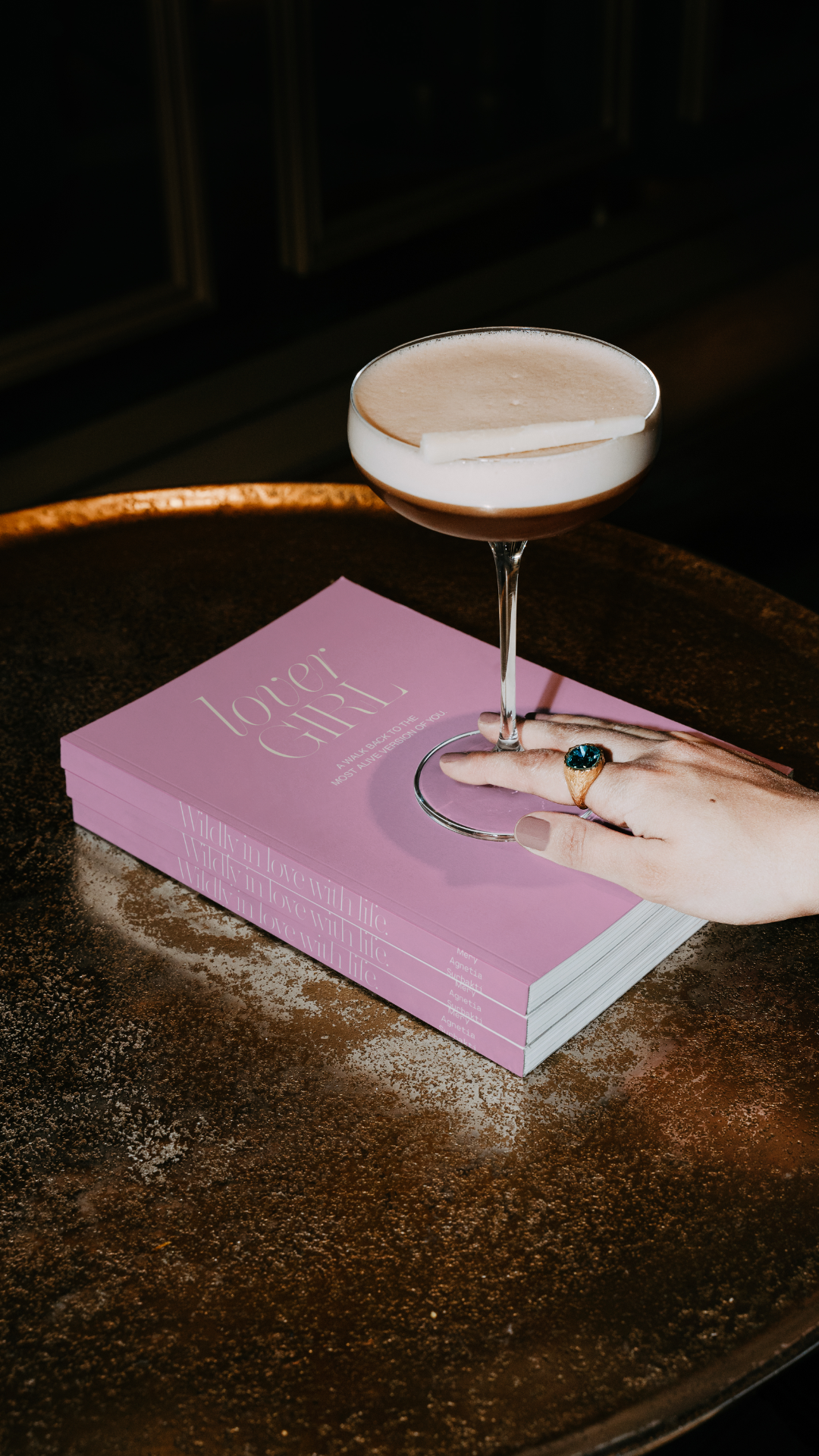 Een hand met een ring die op een roze boek ligt, met daarop een cocktailglas en een boekenrapport.