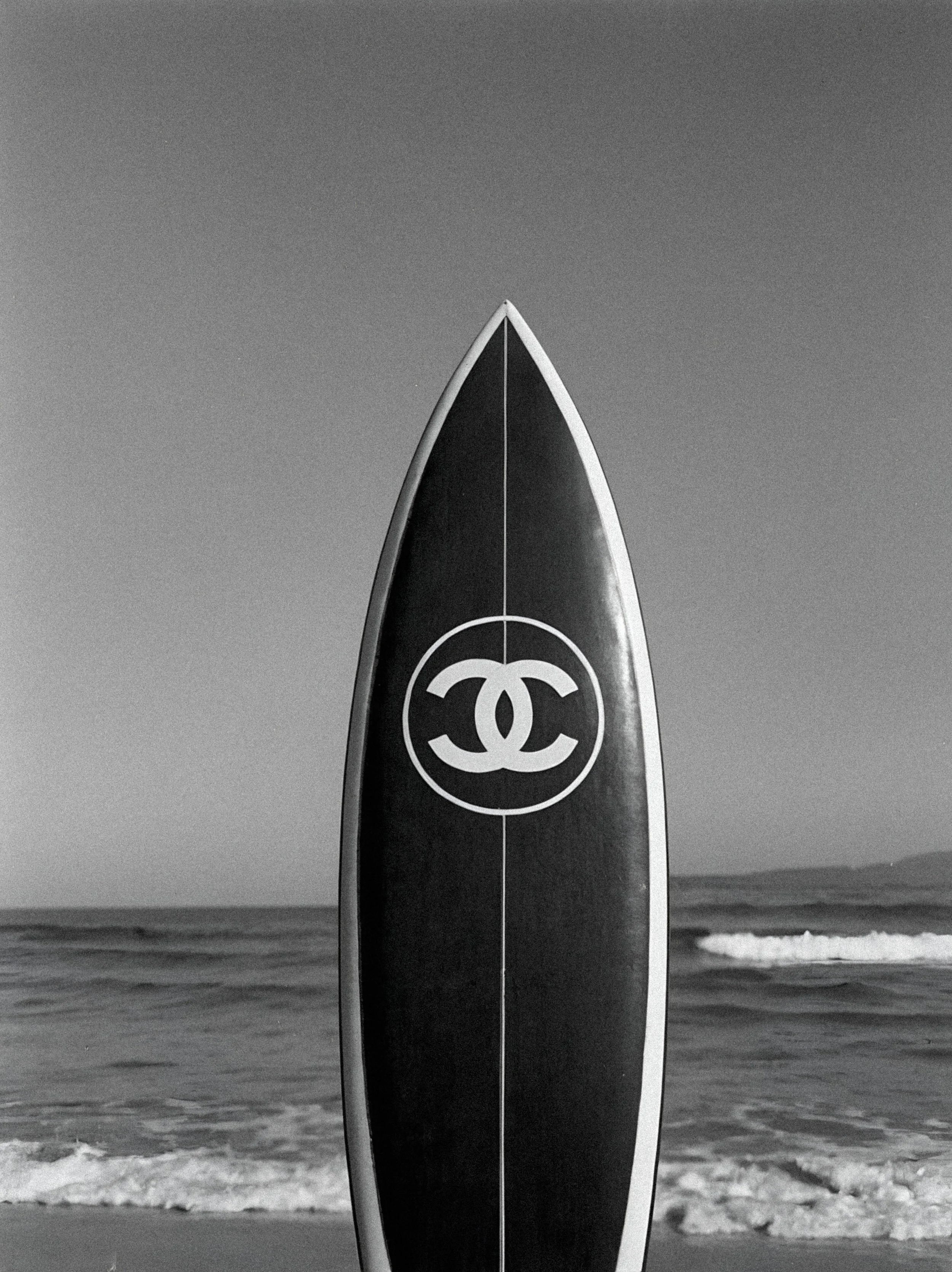 Zwart-wit foto van een surfplank met een Chanel-logo, overeind in de branding van de oceaan met golven op de achtergrond.