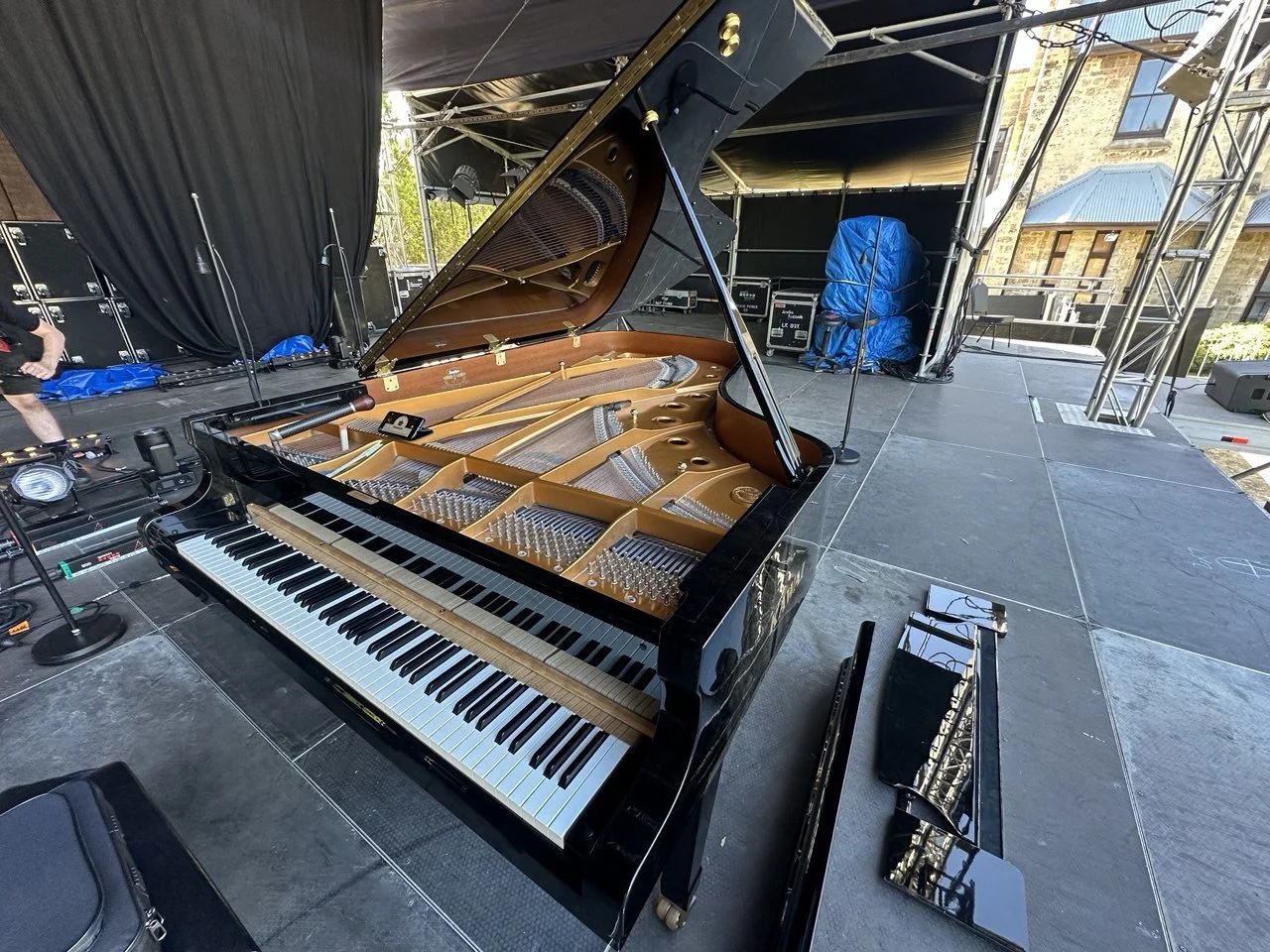 Grand piano on stage with open lid, set up for a performance, with stage equipment and black curtains in the background.