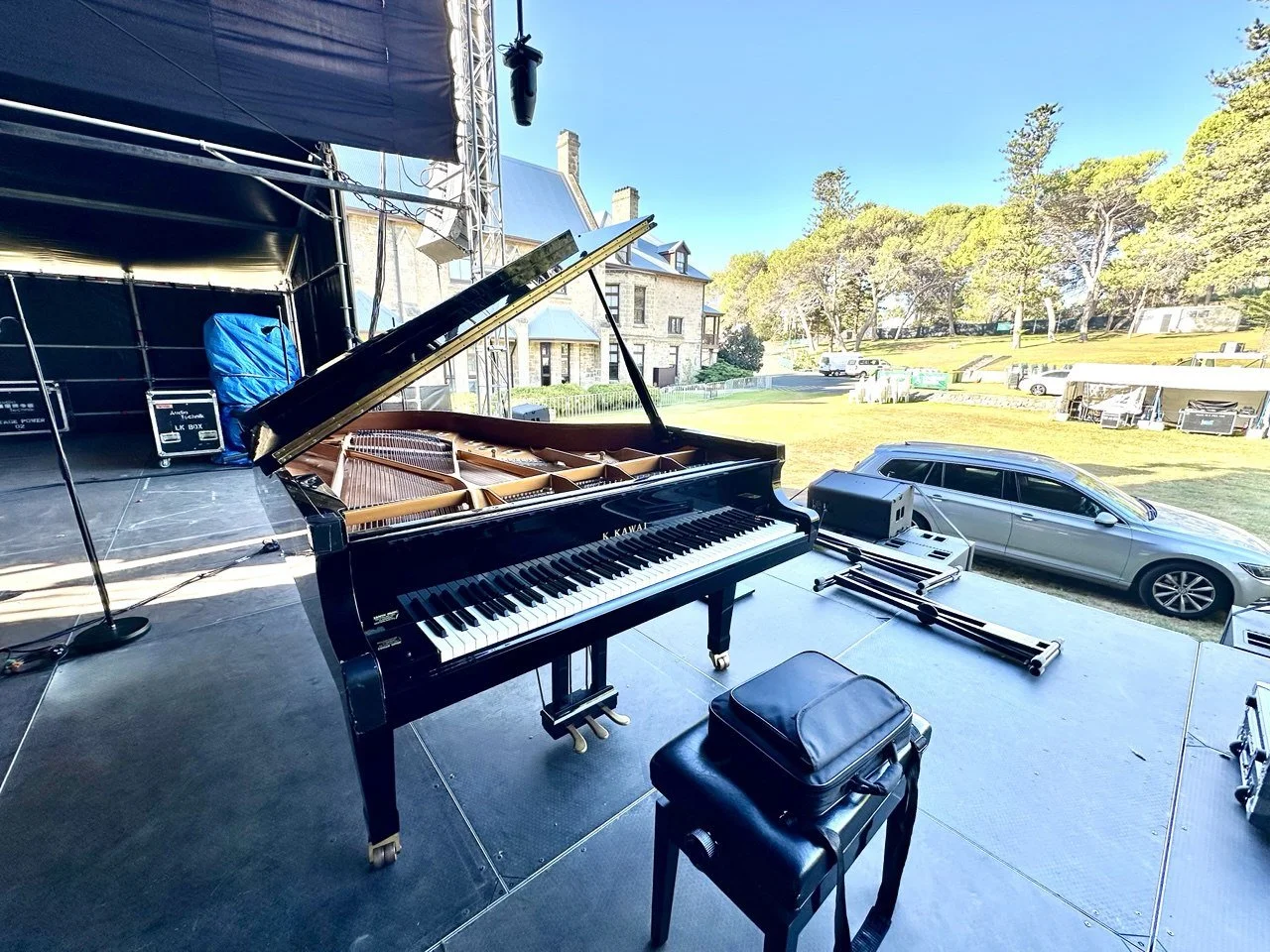 A grand piano on an outdoor stage with its lid open, surrounded by equipment and a chair, overlooking a grassy outdoor area with trees and parked cars.