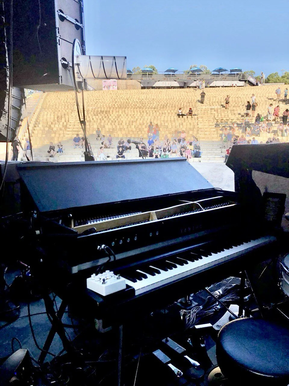 View from stage showing a black grand piano and pedal, with the outdoor amphitheater filled with people sitting on steps in front of the stage, under a clear blue sky.