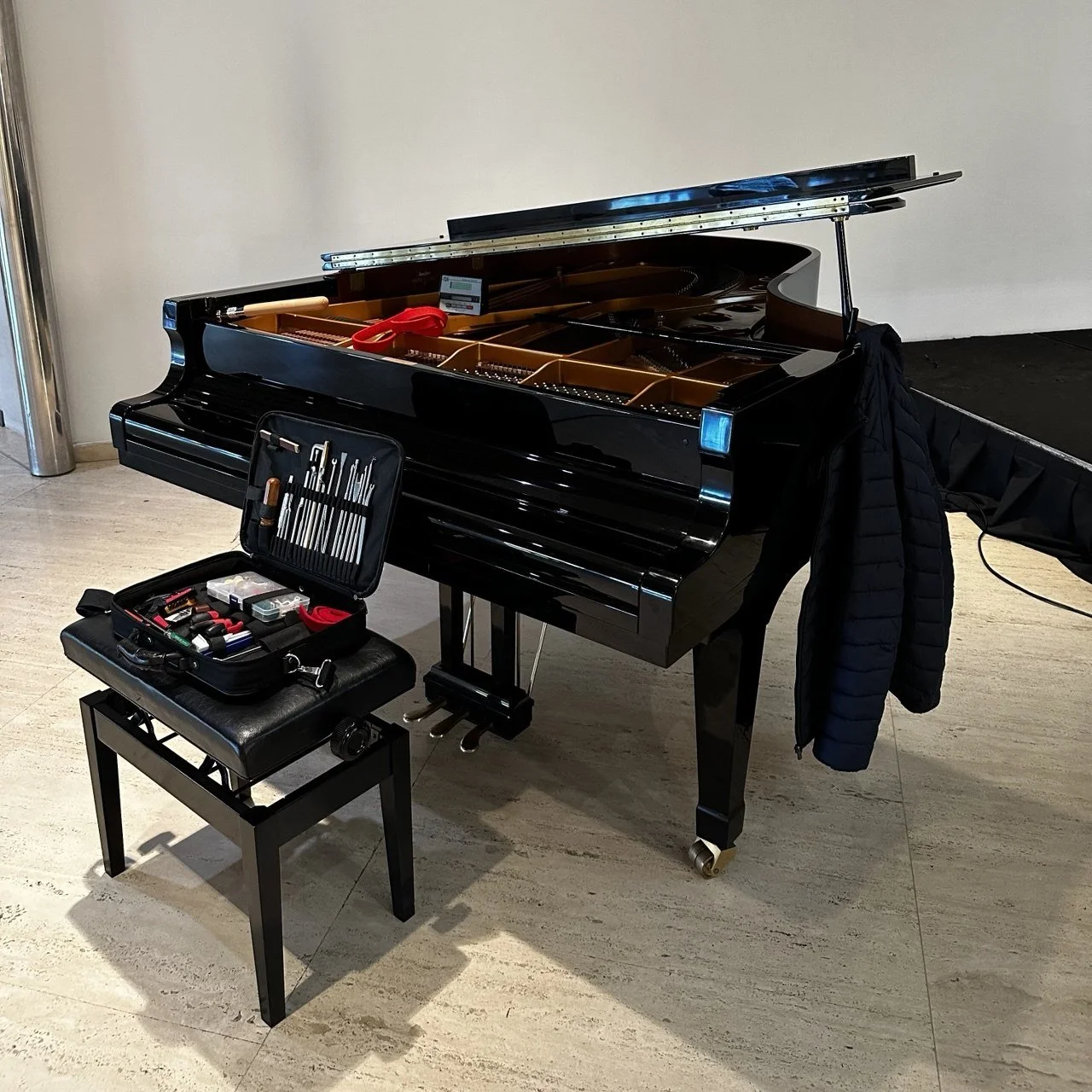 A glossy black grand piano with its lid open, showing the strings and interior, is accompanied by a small bench with a tool kit and maintenance supplies on top. A black puffy coat hangs on the side of the piano, and the setup appears to be for tuning or maintenance.
