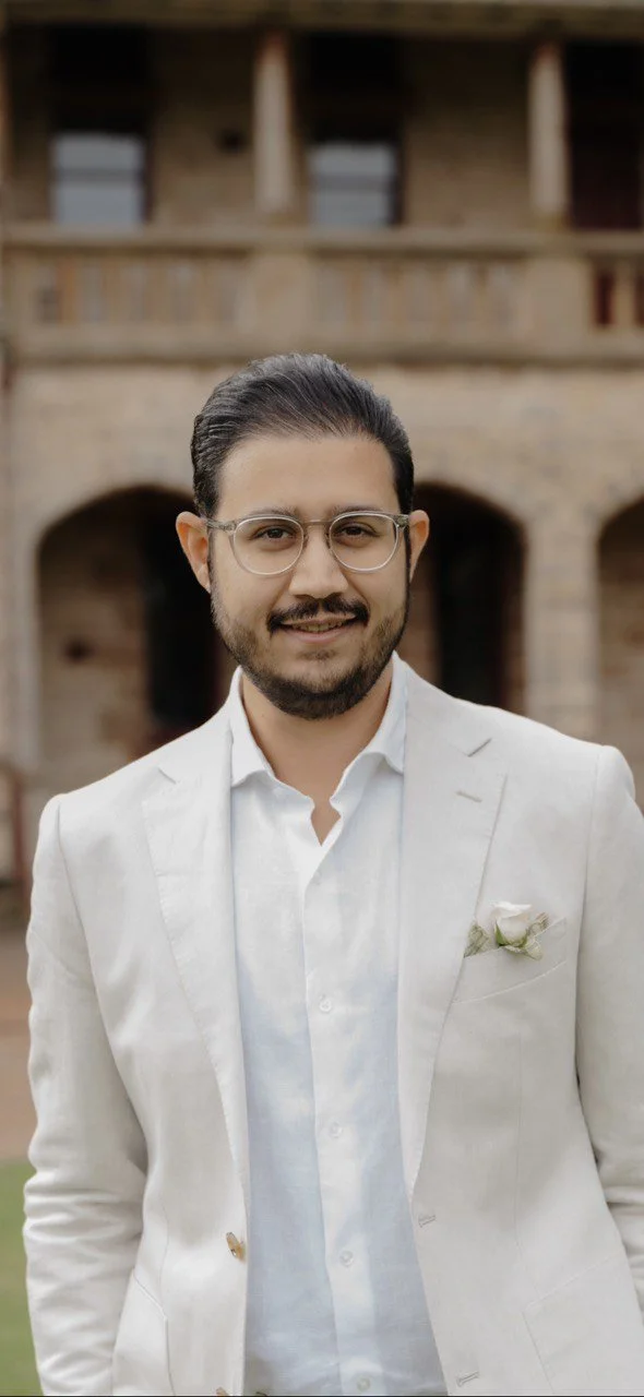 A man with dark hair, glasses, and a beard, dressed in a white suit with a flower lapel pin, standing in front of a historical brick building.