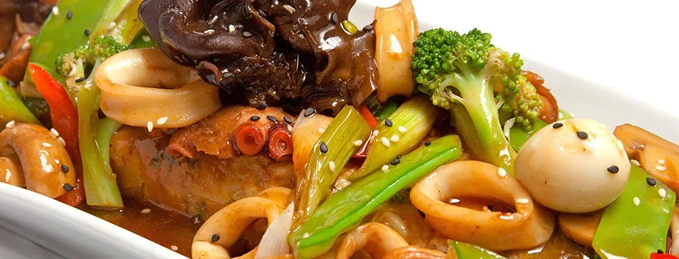 Close-up of a bowl of stir-fried vegetables and seafood, including broccoli, quail eggs, squid, wood ear mushrooms, and sliced carrots, garnished with sesame and black seeds.