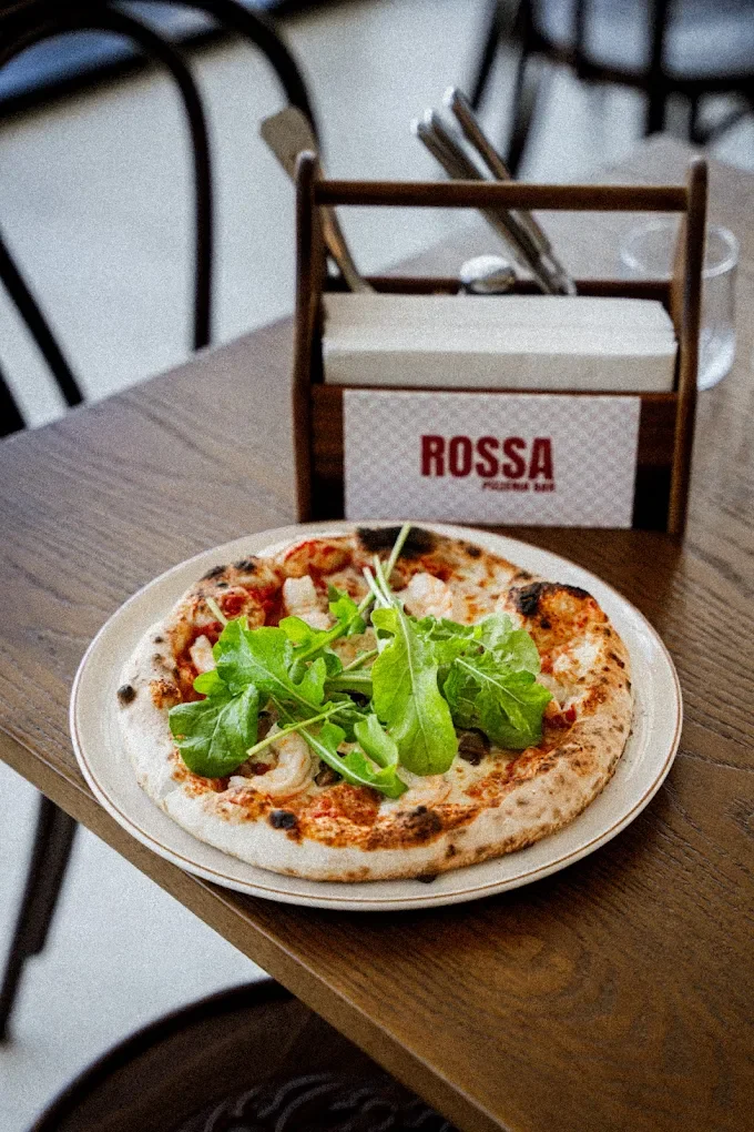 A pizza with cheese and toppings topped with fresh greens, on a white plate on a wooden table.