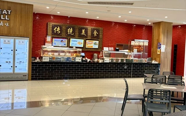 Modern shopfront with shiny lit up bain marie's glass fridges with fresh food to the left of the counter, large gold chinese writing on a black framed with wood background, with red wall behind.