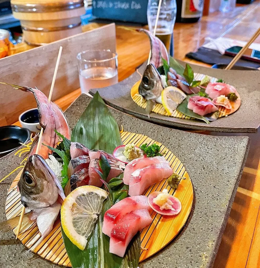 Two plates of sashimi featuring fish heads and slices of raw fish, garnished with lemon slices, wasabi, and green herbs, on a table in a restaurant.