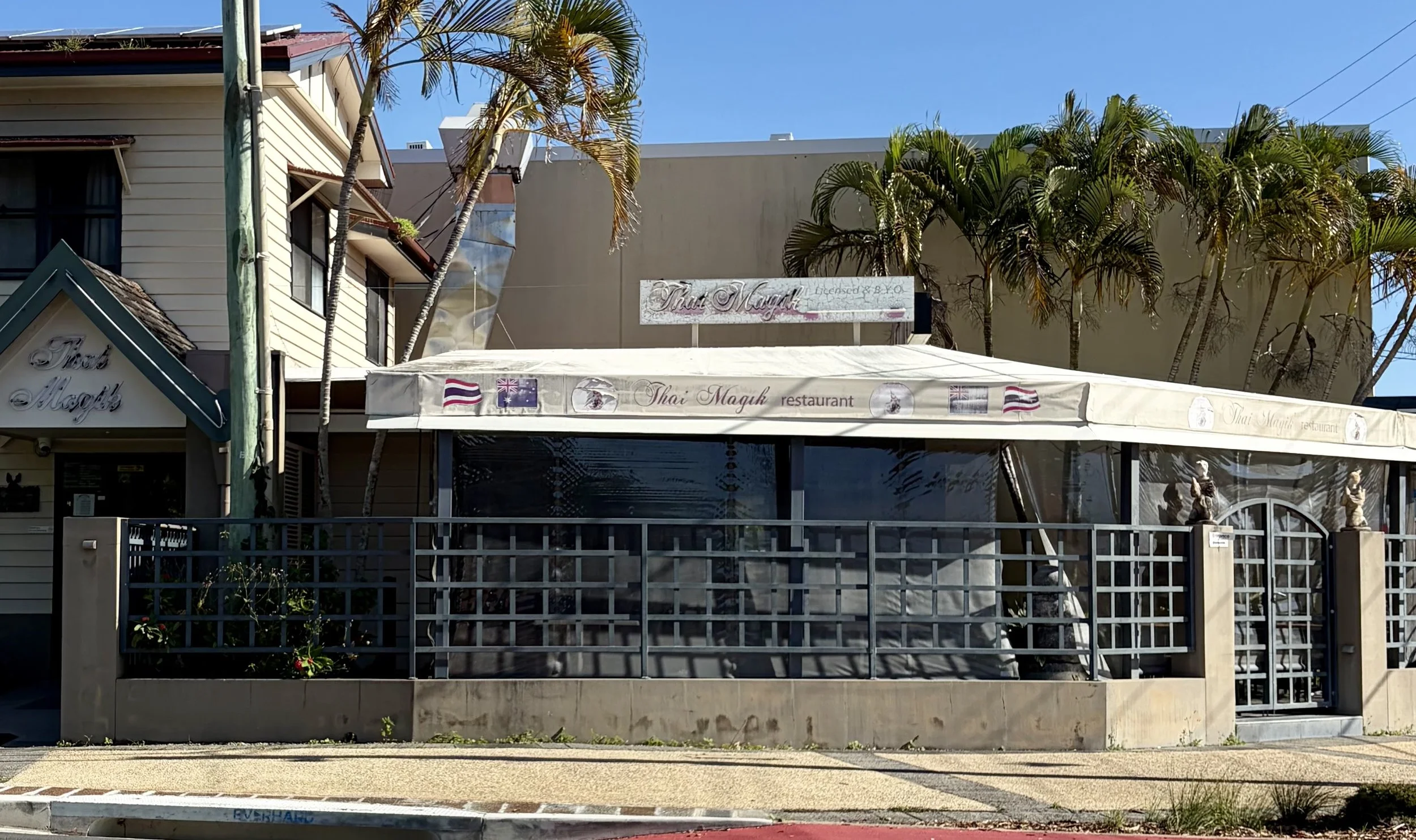 Exterior of a Thai restaurant named Thai Maguy, with a canopy and outdoor seating, surrounded by a small garden and palm trees, on a street with traffic and a no cycling sign.