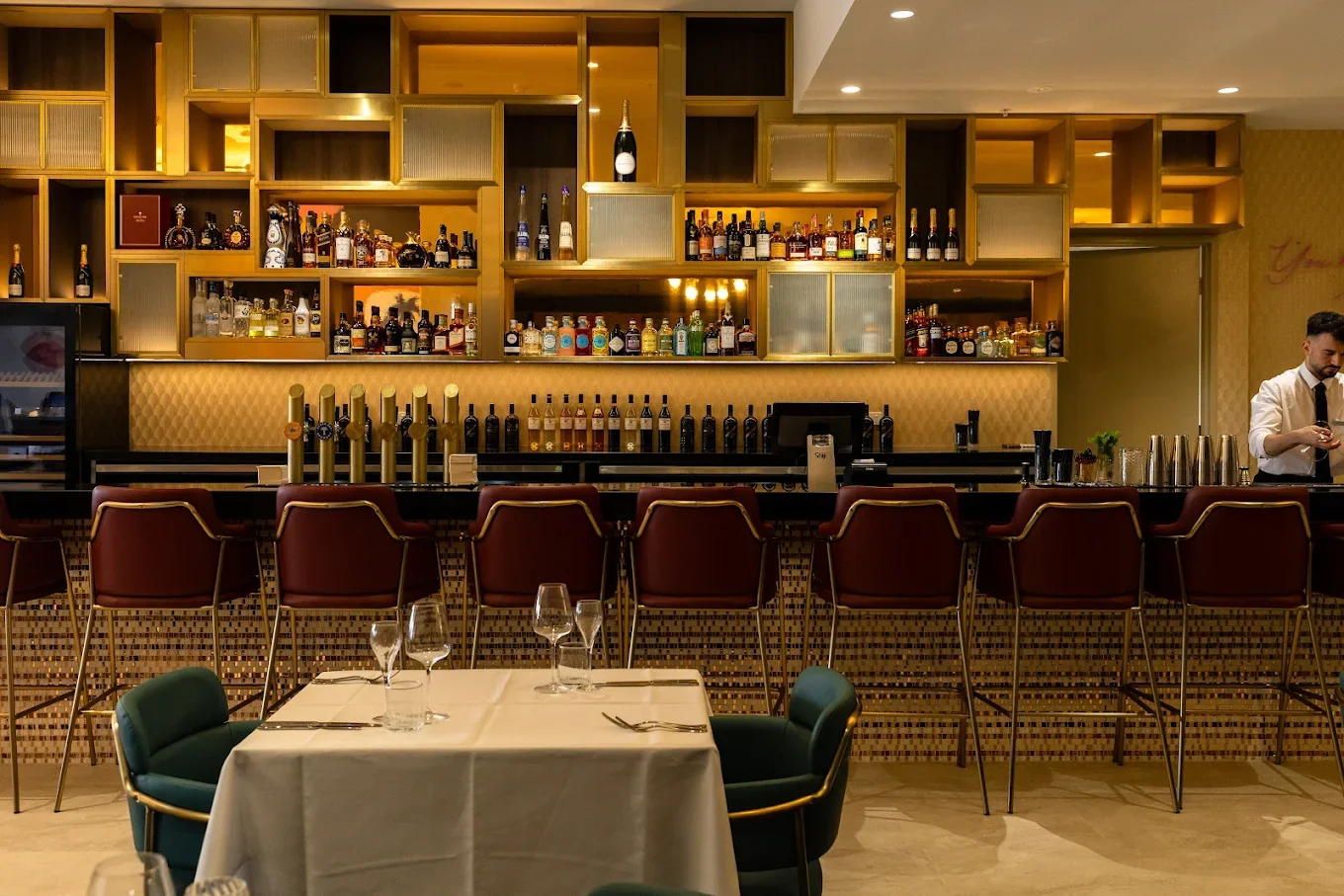 Interior of a modern bar with a tiled front counter, bar stools, and a back wall with shelves full of liquor bottles, a table set for dining, and a bartender on the right using a mobile phone.